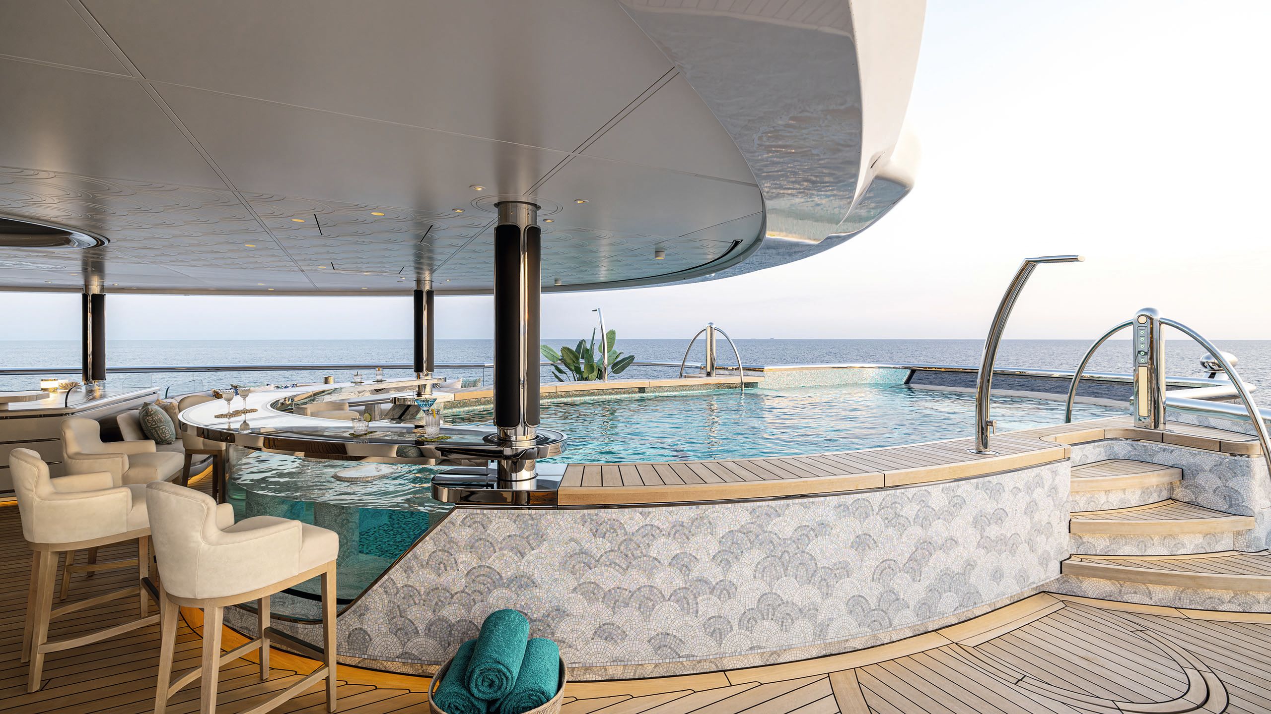 A curved pool with mosaic tiling and see-through side that doubles as a bartop. There are high chairs all around and two martini glasses