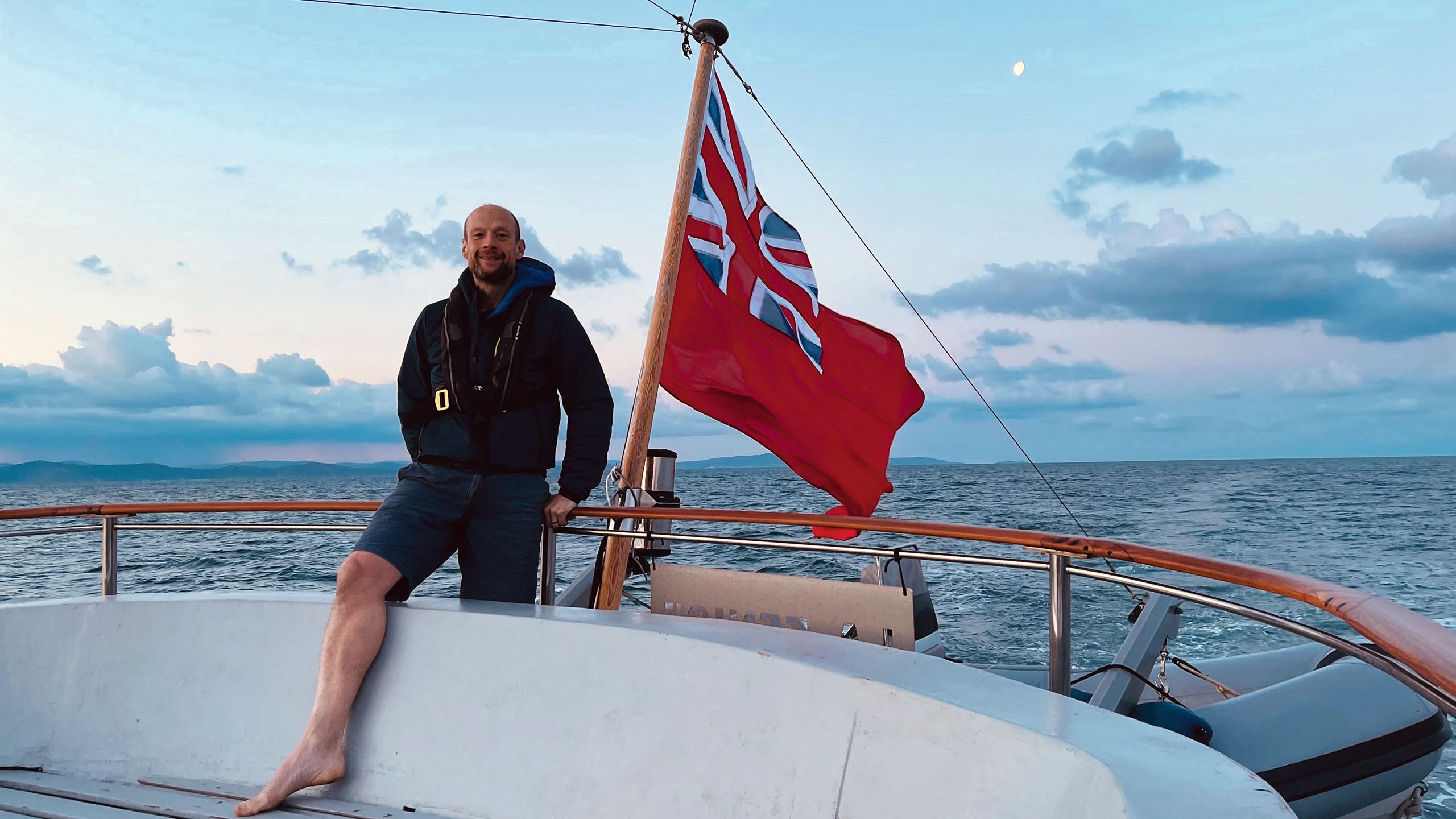 Leigh Shardlow standing on the edge of his boat, which has a Red Ensign flag at the end