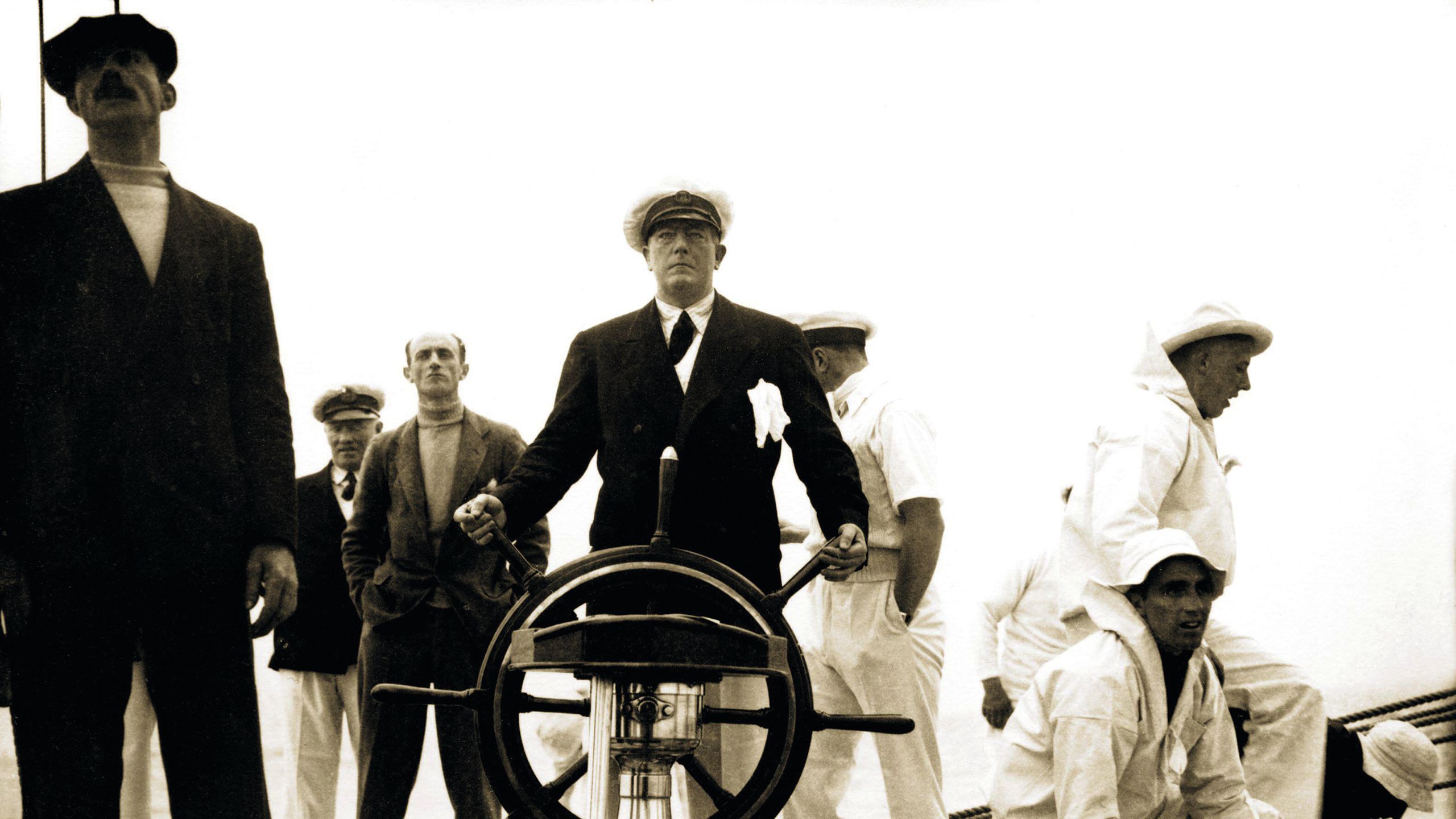 A sepia photo of Sir Richard Fairey, surrounded by other men, some in white sailor outfits,steering a vessel
