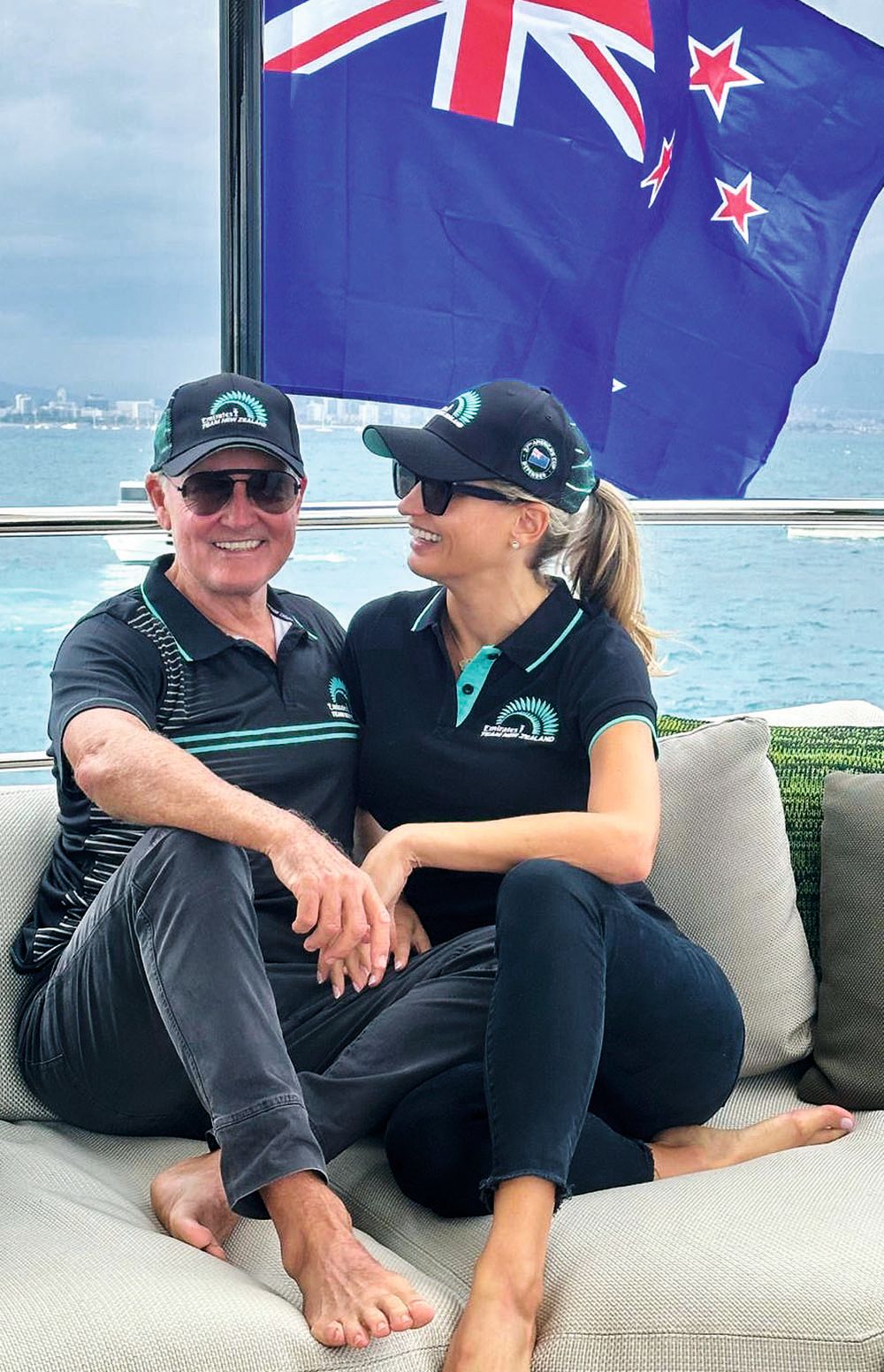 Owen and his partner Madeleine are wearing dark blue polo tops and trousers with matching caps and sunglasses. She is turned to him and smiling and he's looking at the camera. A New Zealand flag is behind them. 