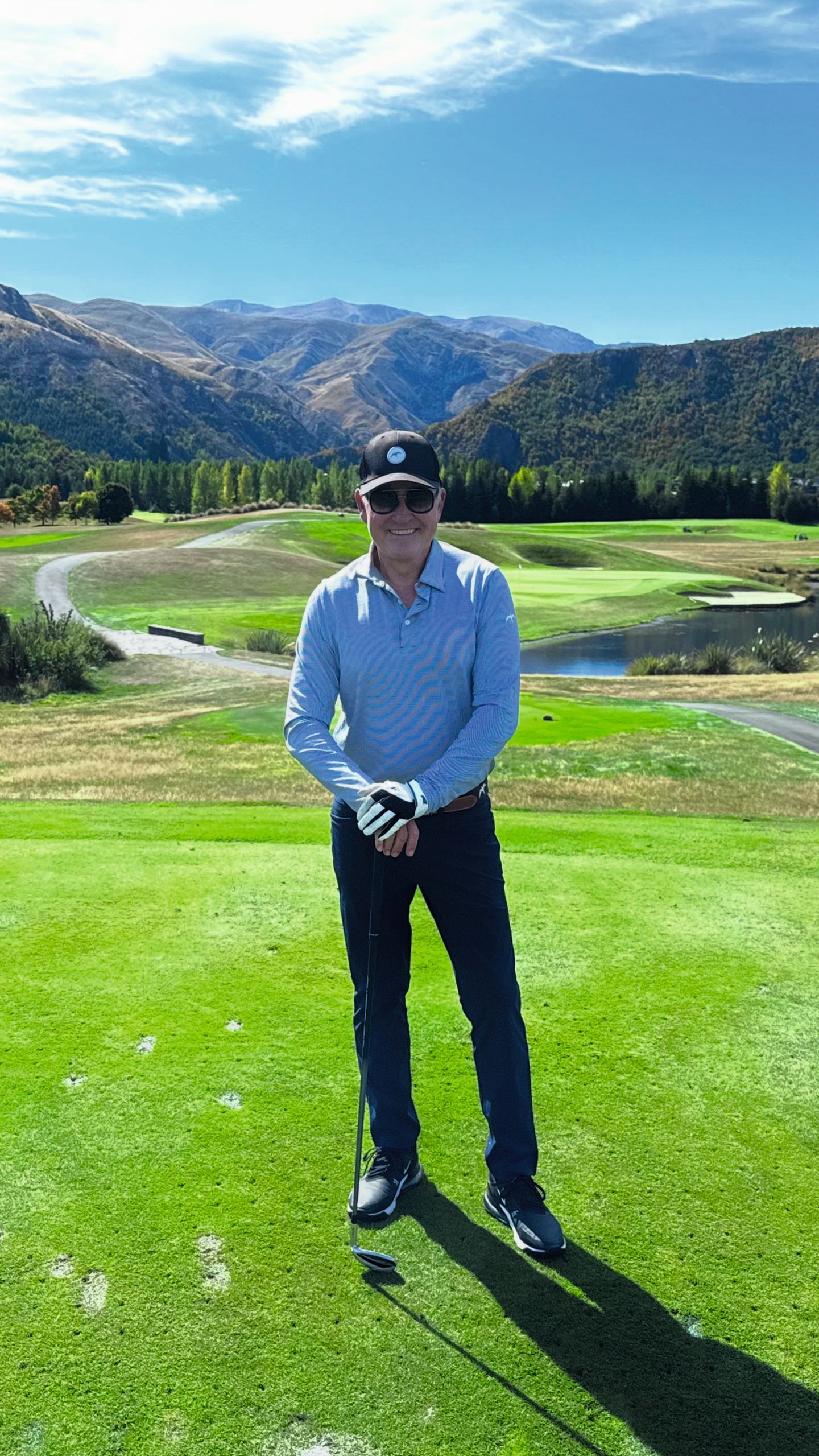 Owen on a golf green. He's wearing dark trousers, a light blue shirt and dark blue cap. He's holding a golf club