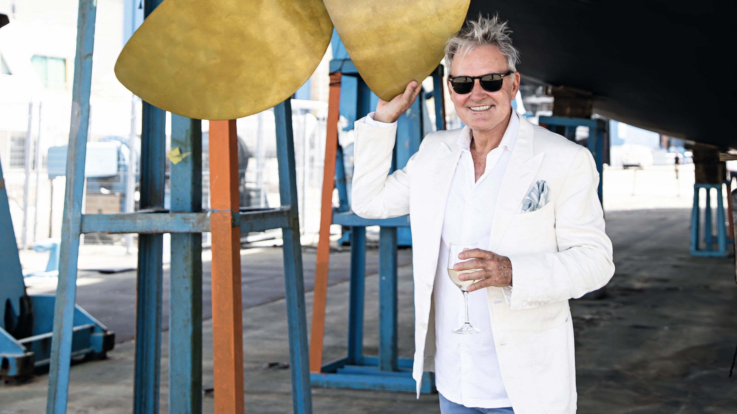 Steve Owea wearing a white suit and black sunglasses, holding a glass of white wine