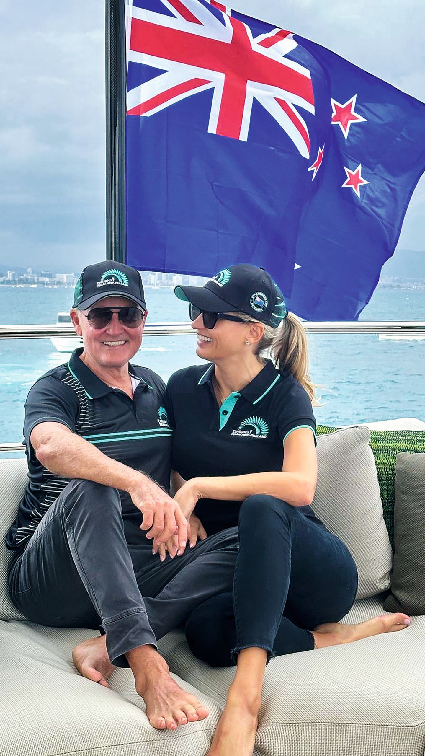 Owen and his partner Madeleine are wearing dark blue polo tops and trousers with matching caps and sunglasses. She is turned to him and smiling and he's looking at the camera. A New Zealand flag is behind them. 