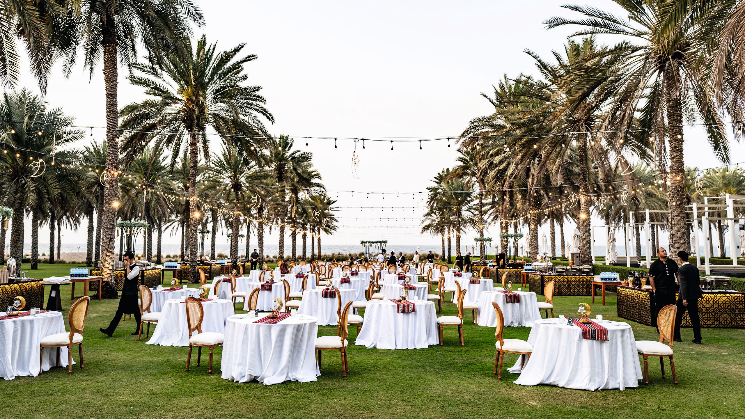 A lawn filled with circular tables covered in white tablecloths. They are lined either side by an avenue of palm trees and festoon lights