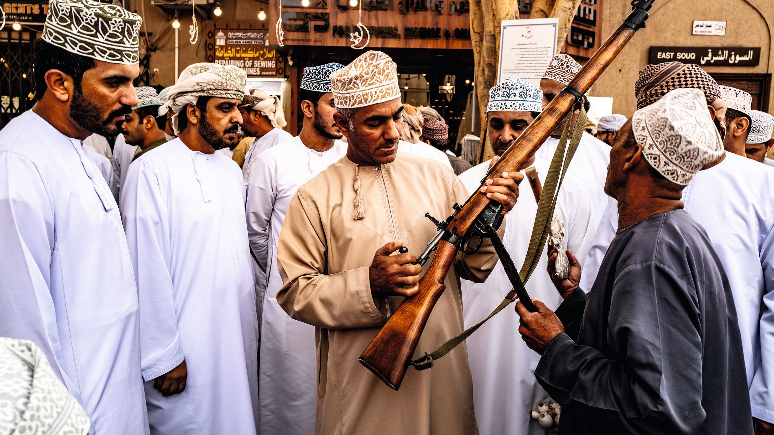 A group of men standing in a huddle, the man in the centre is holding a large shotgun/rifle