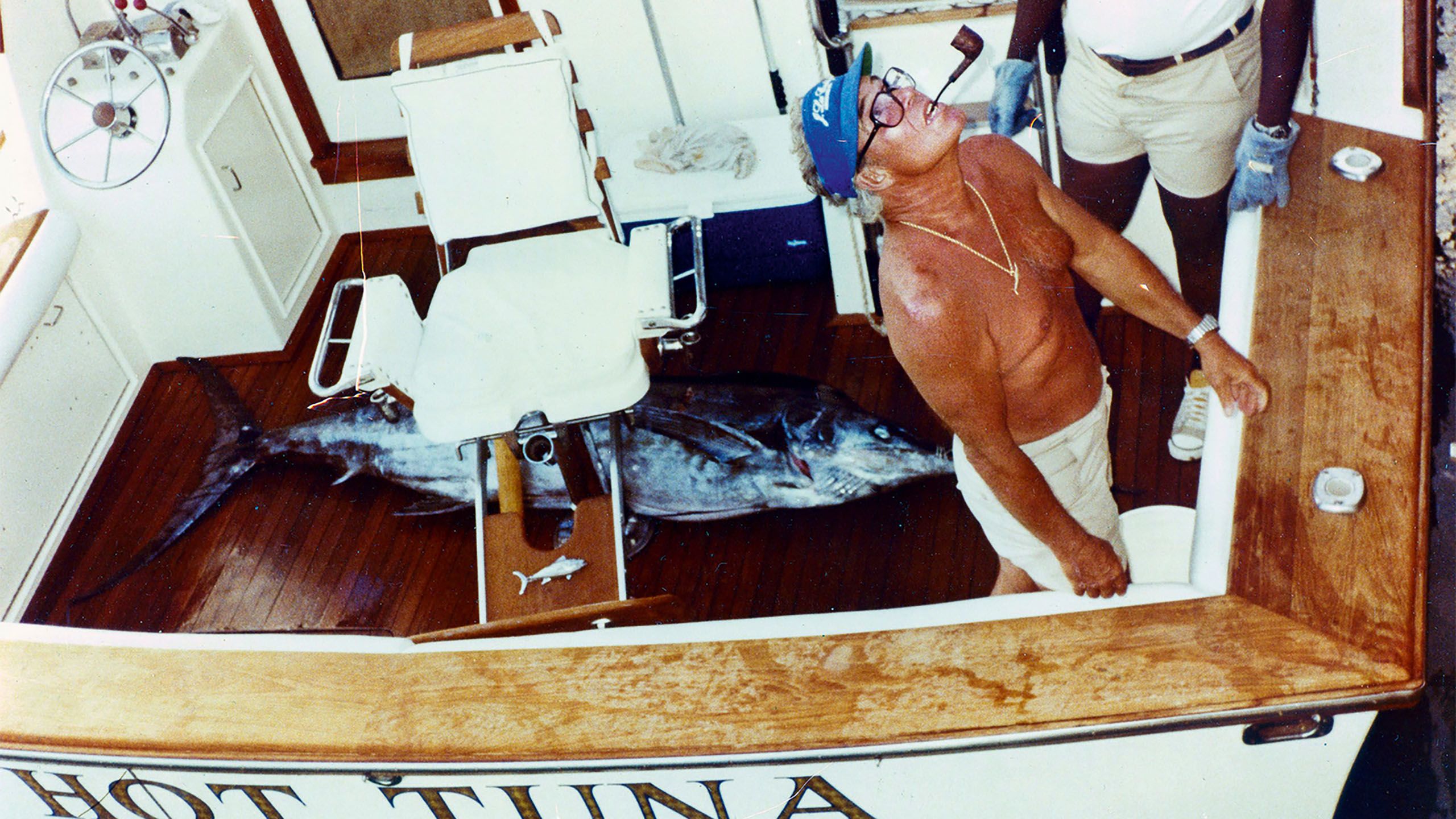 A shirtless man with a very deep tan, wearing black rummed glasses, a blue cap and a pipe sticking out of his mouth. He is on a boat called Hot Tuna