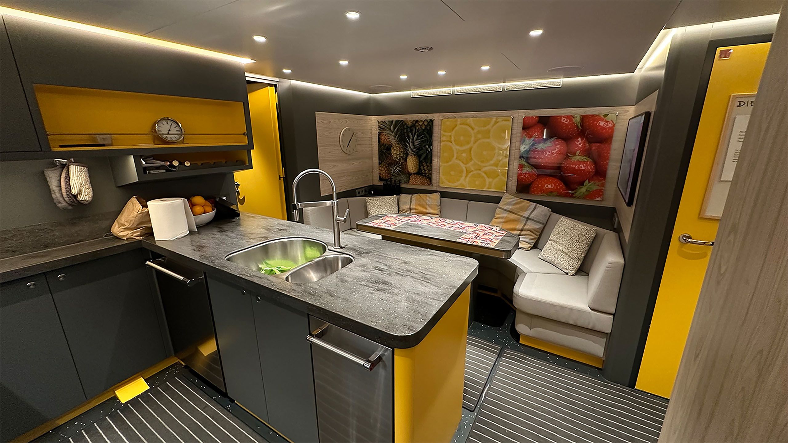 Interior view of a small kitchen with grey and yellow counters and cupboards. Opposite is curved banquette seating around a table
