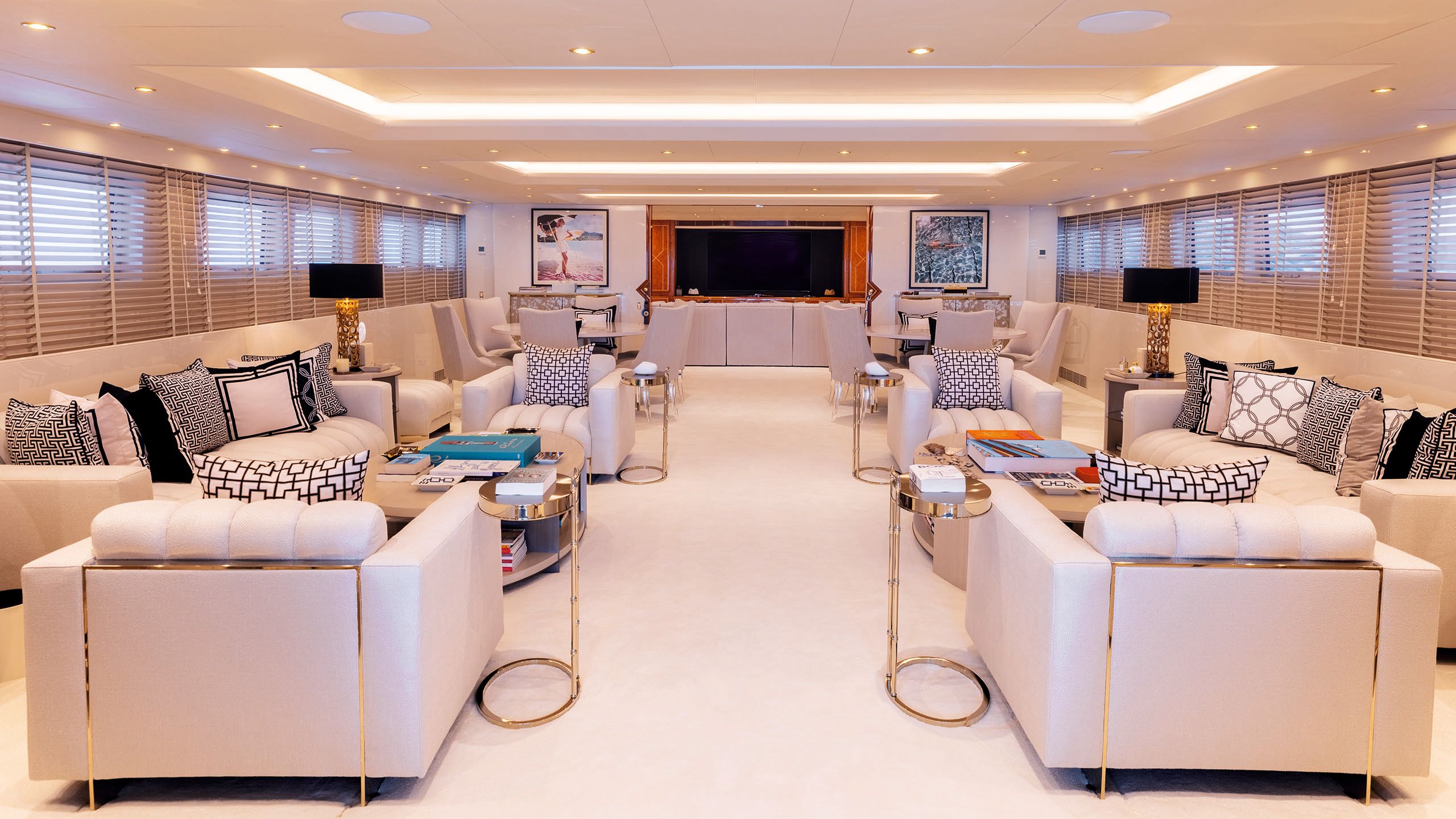 Wide shot of the interior living space. There are two large sofas facing each other and two armchairs in the foreground. The seating is a cream colour and the cushions are monochrome with geometric patterns. There are gold side tables by the arm chairs and large oval coffee tables in front of the sofas