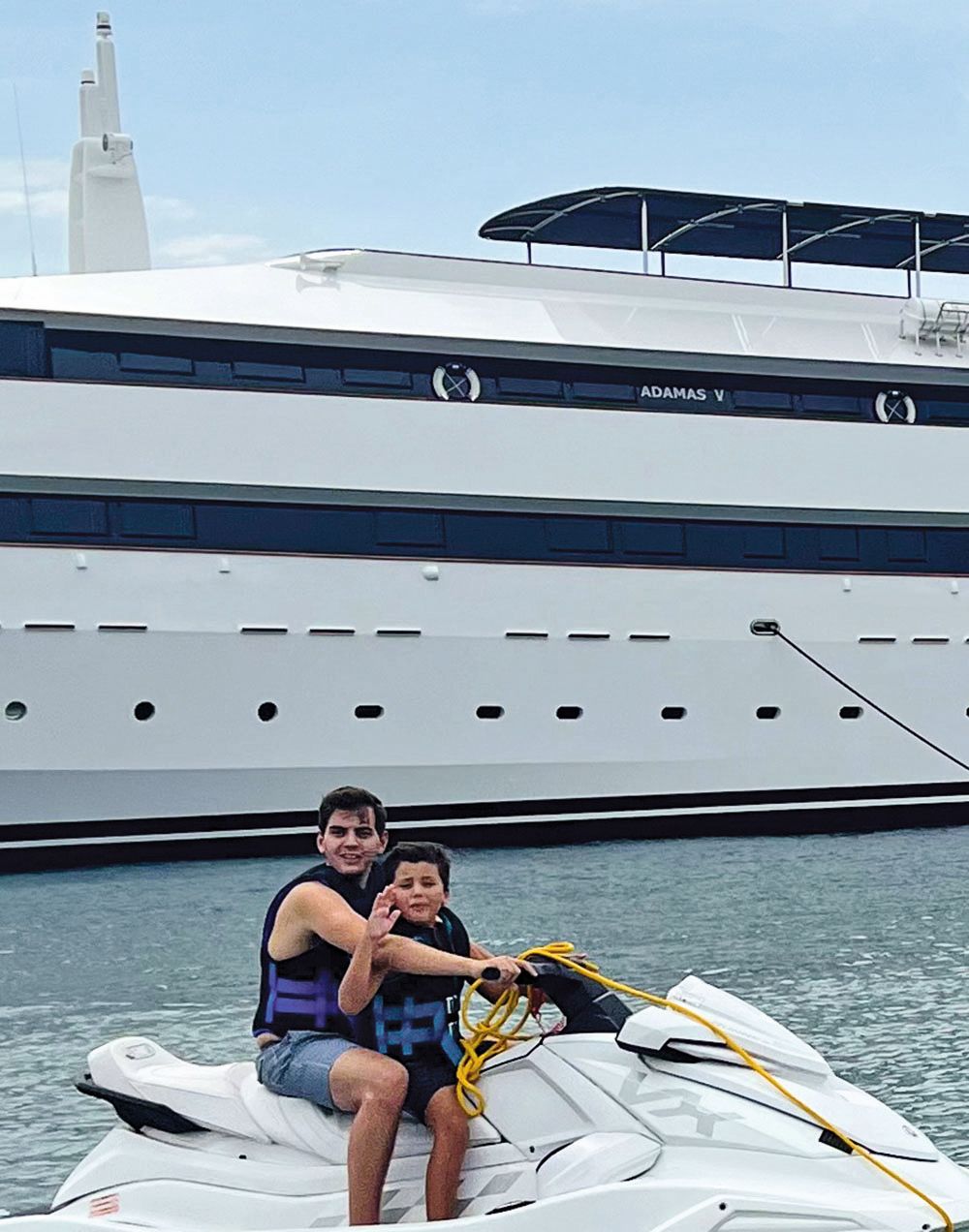 Two young boys in life vests sitting astride a jet ski with a large white boat in the background