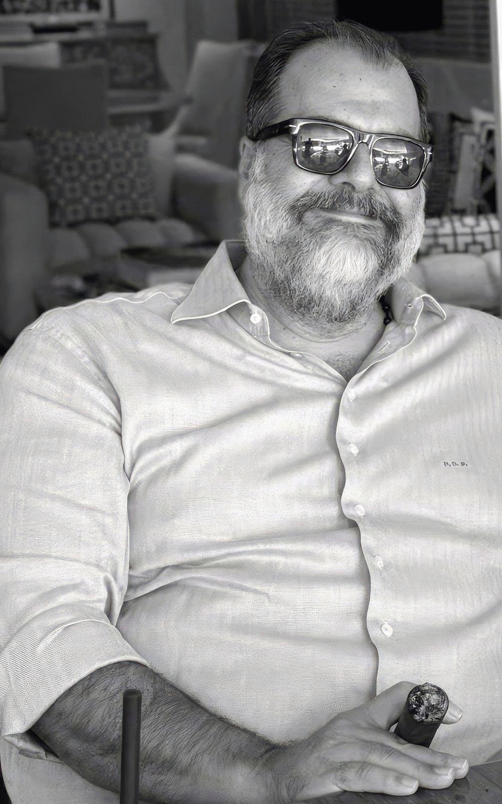 A black and white photo of Pateras. He is wearing sunglasses and a light shirt and is smiling at the camera