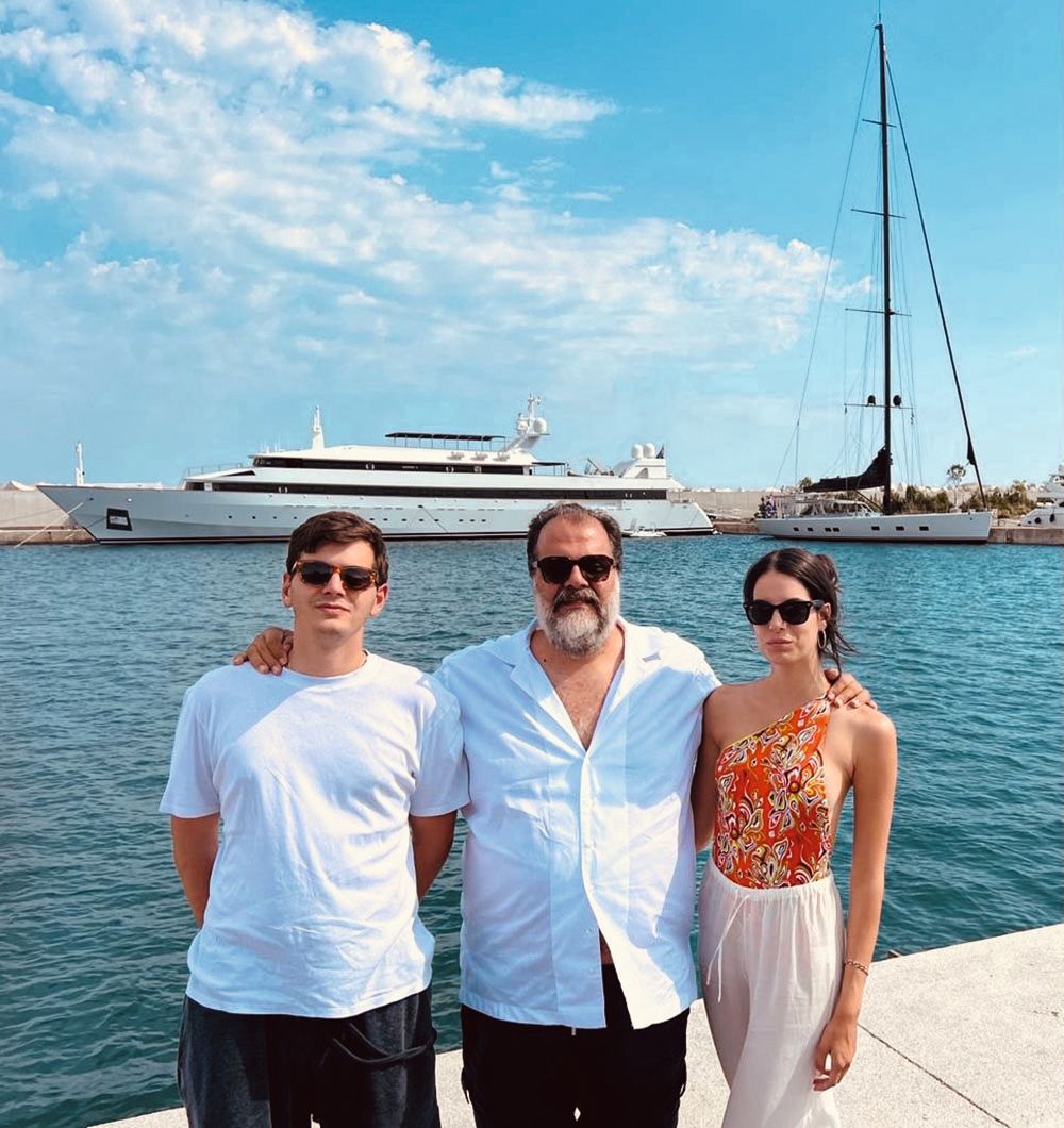 Pateras is in the centre with his arm around his son to the left and daughter to the right. He is wearing a light-coloured, casual shirt, his son is wearing a white t-shirt and his daughter is wearing white linen trousers and an orange, patterned swimsuit. They are all wearing sunglasses. Their vessels are in the background