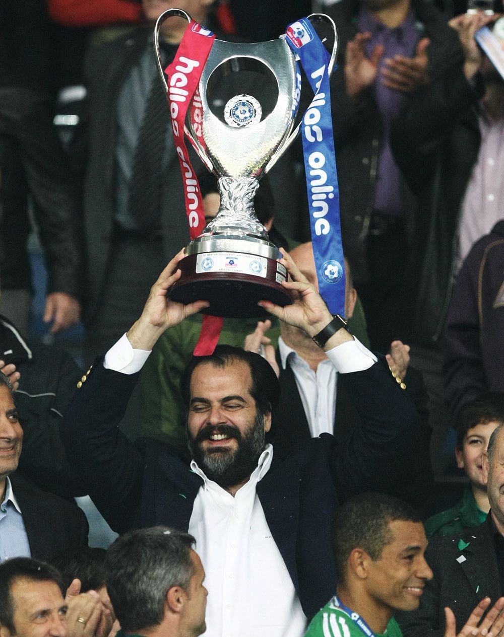 Pateras in a black suit jacket and white shirt, holding a silver trophy aloft which has a blue ribbon on one handle and a red on the other