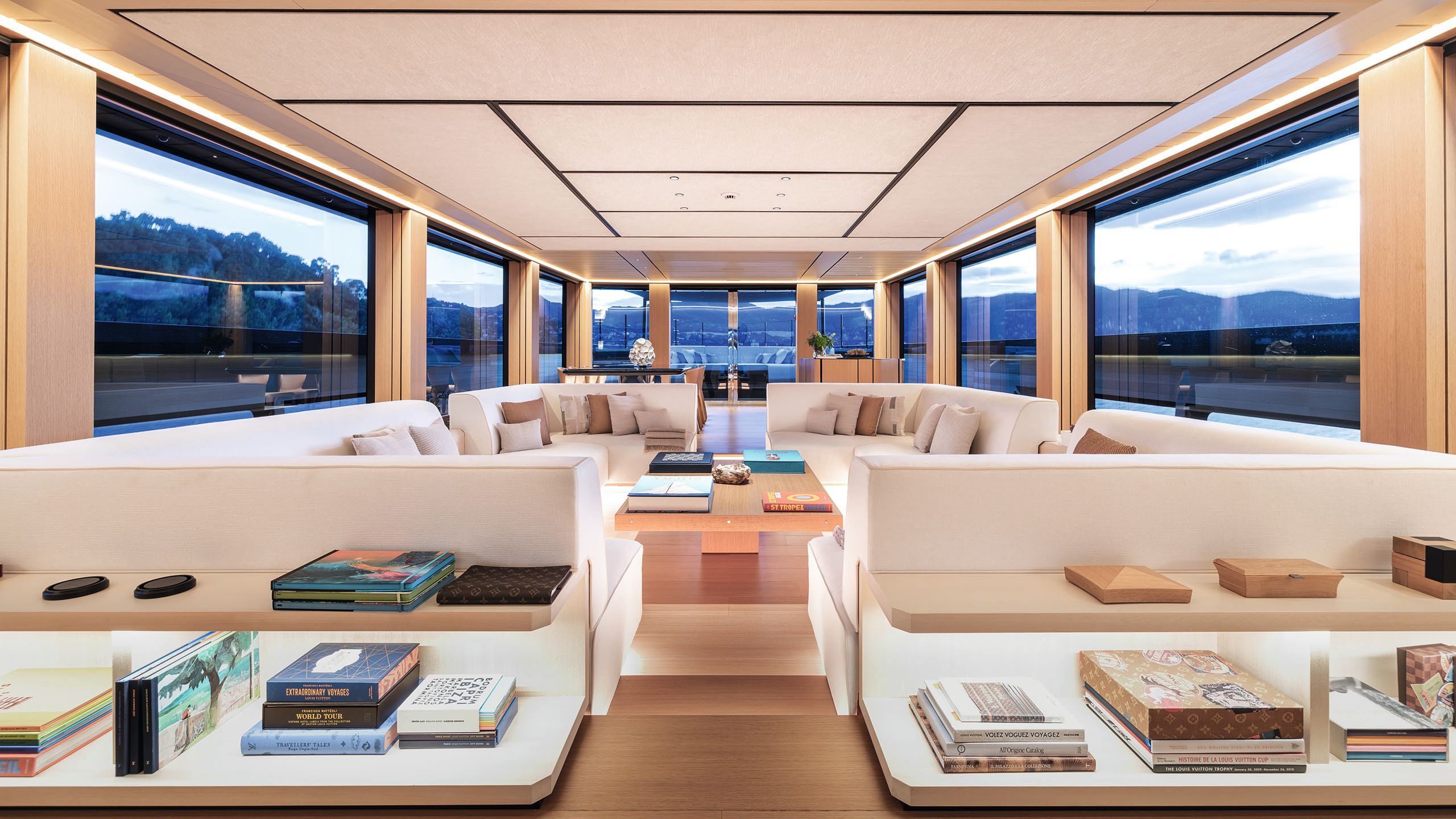 Four L-shaped white sofa arranged in a square with a table in the centre. In the foreground are shelves with books stacked on top