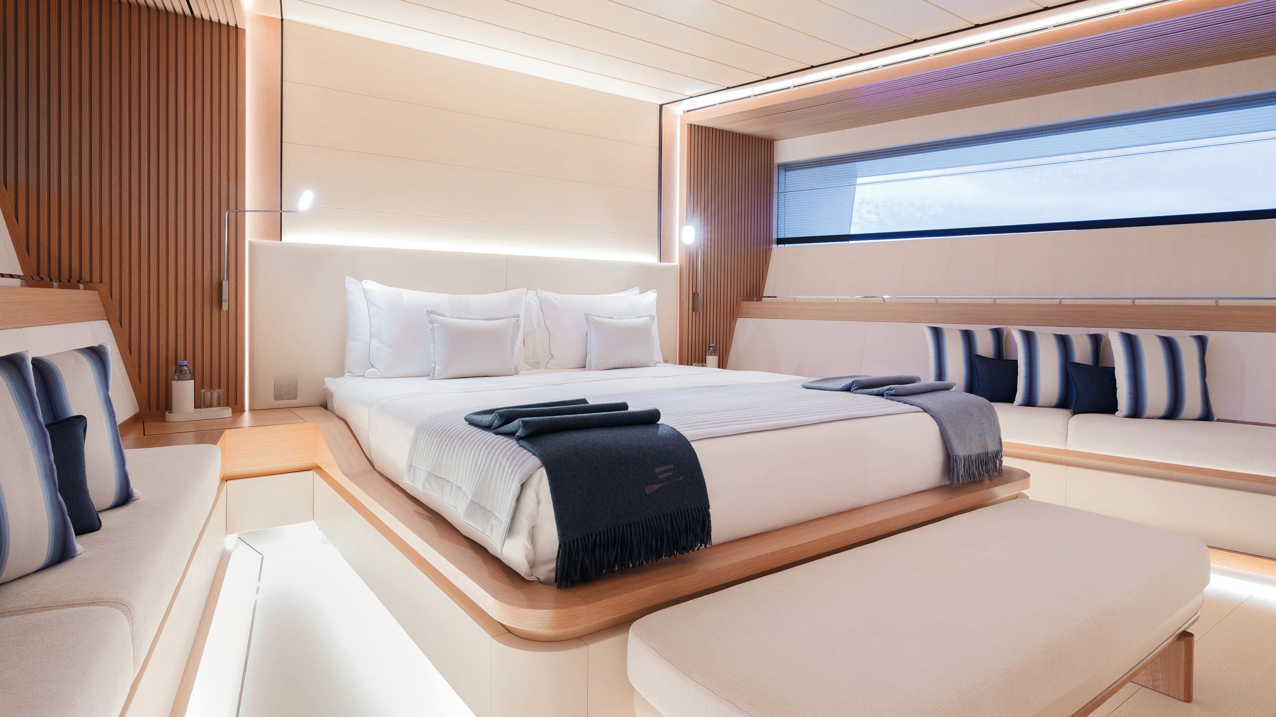 The master bedroom has a bed made up with white linens and blue throws. On either side of the bed is banquette seating with blue and white stripe cushions