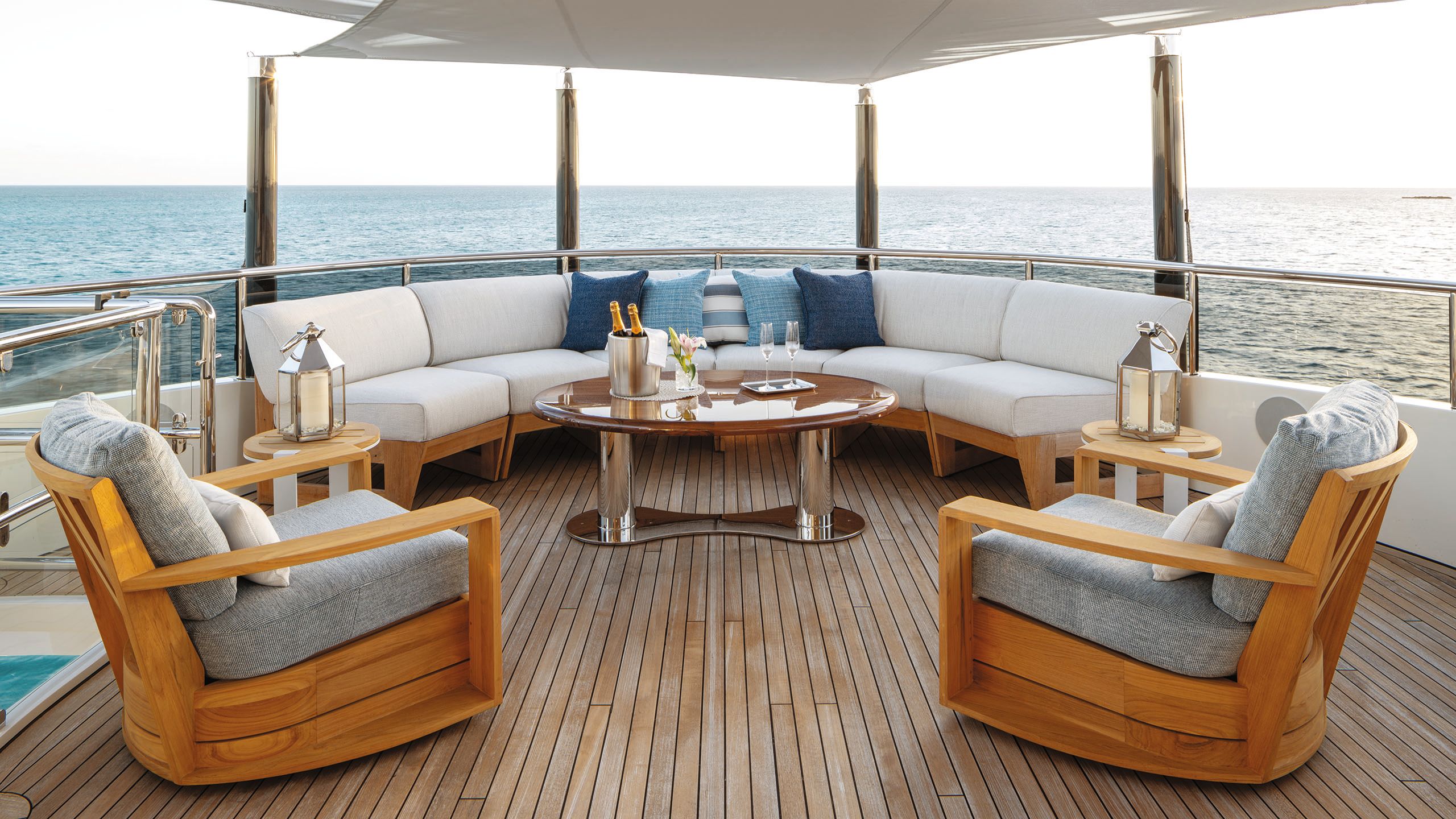 An outdoor deck with teak armchairs and grey cushions and a curved sofa with neutral cushions and an oval table in front