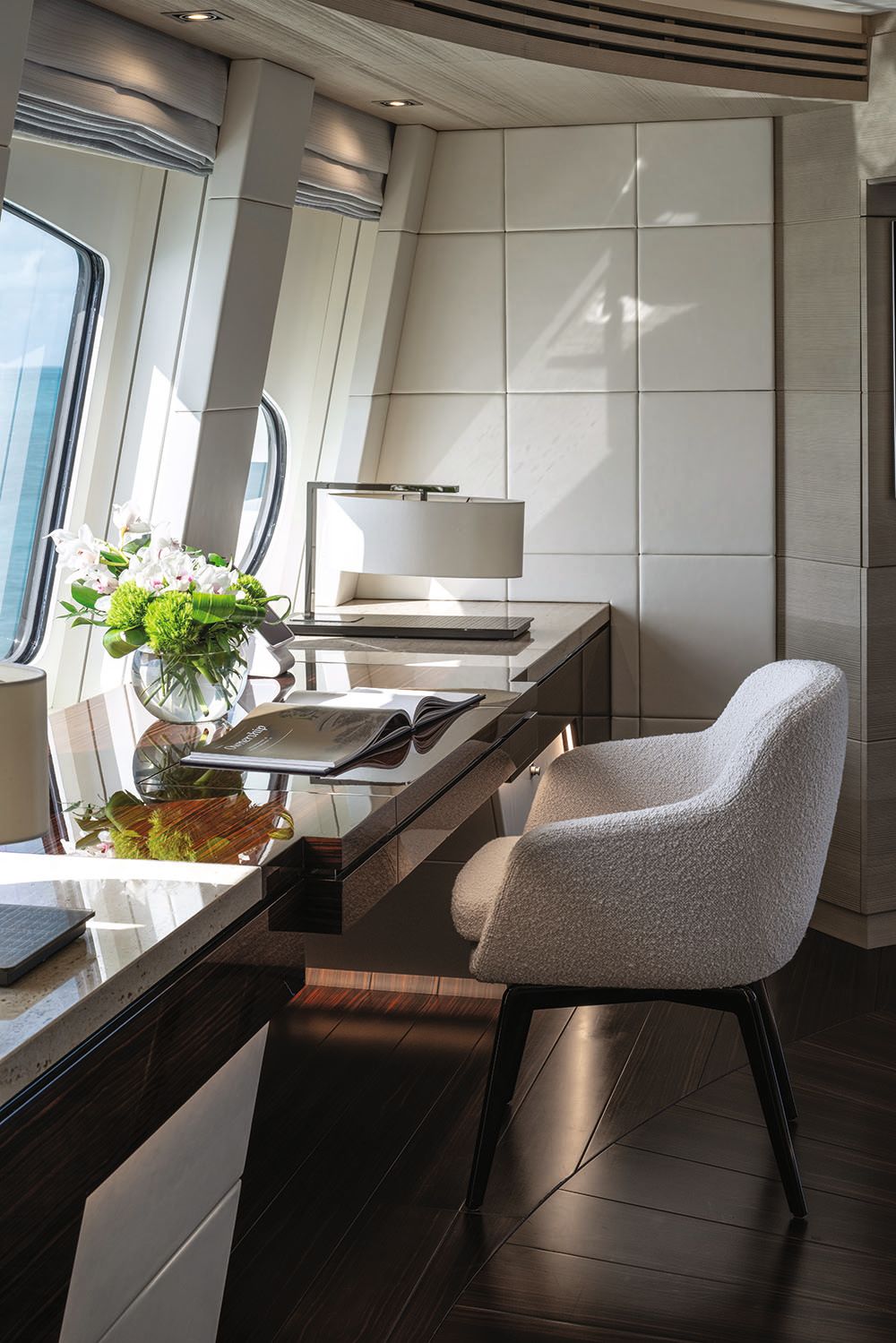 Close-up of a wooden desk with upholstered chair in neutral fabric. There's a base of flowers, a lamp, and a book on the table