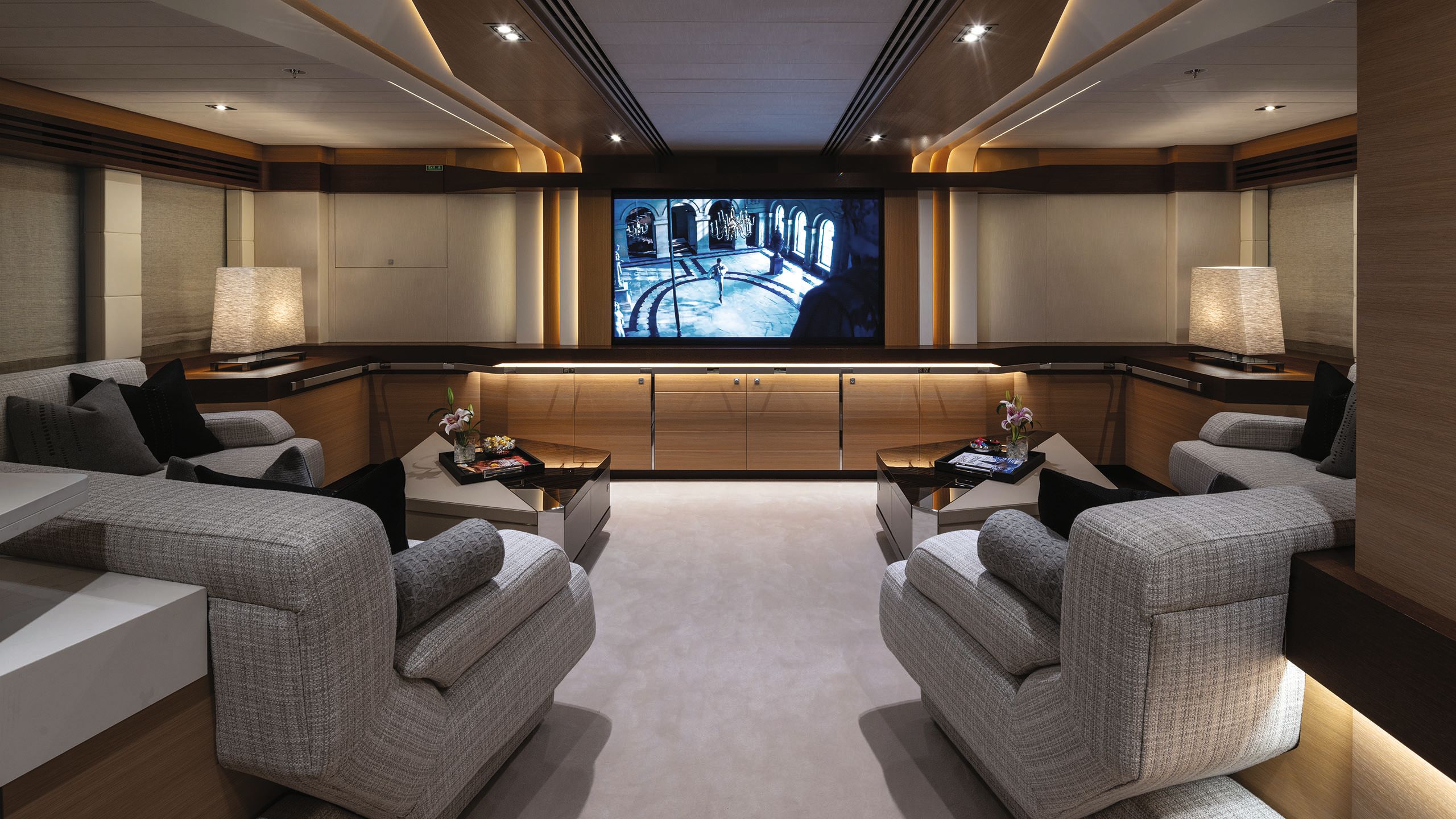 Wide shot of the cinema room, taken from the back. There are large grey sofa seats and a screen on the wall at the back