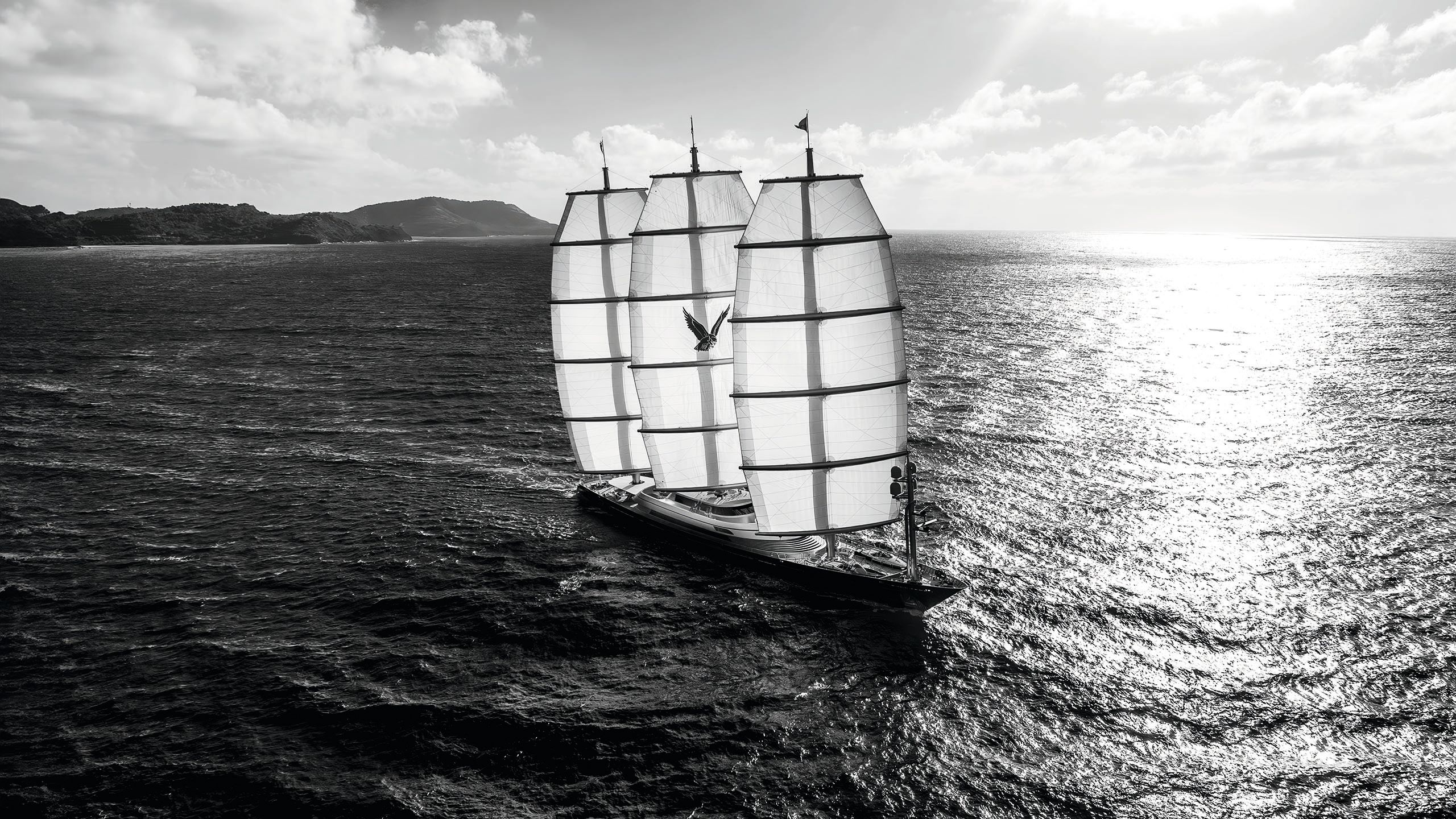 Black and white shot of Maltese Falcon on the water, taken from afar. The sunlight is shining through its three sails