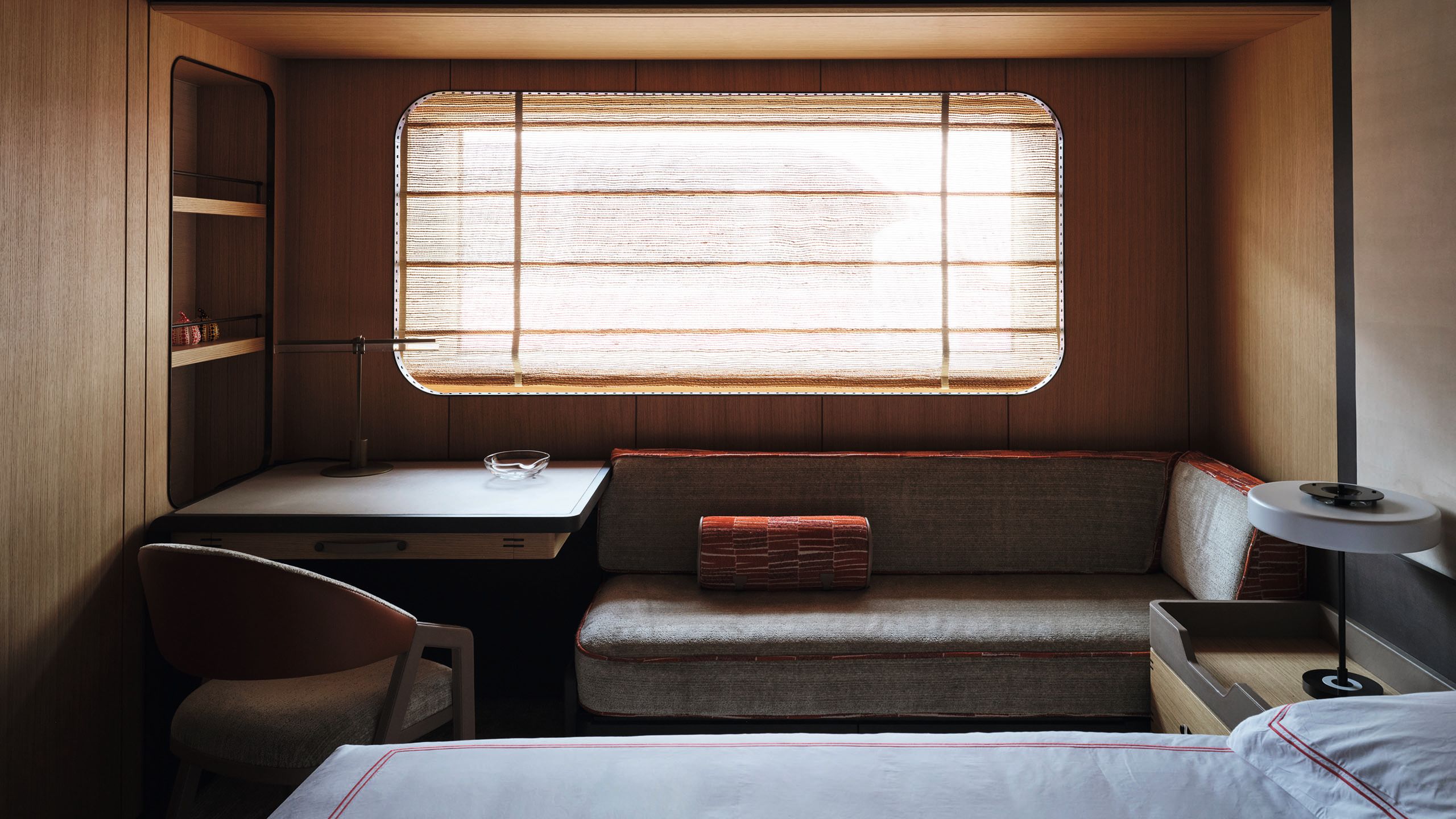 A rectangular window with a blind, above a small table, chair and L-shape sofa. The edge of a bed is in the foreground