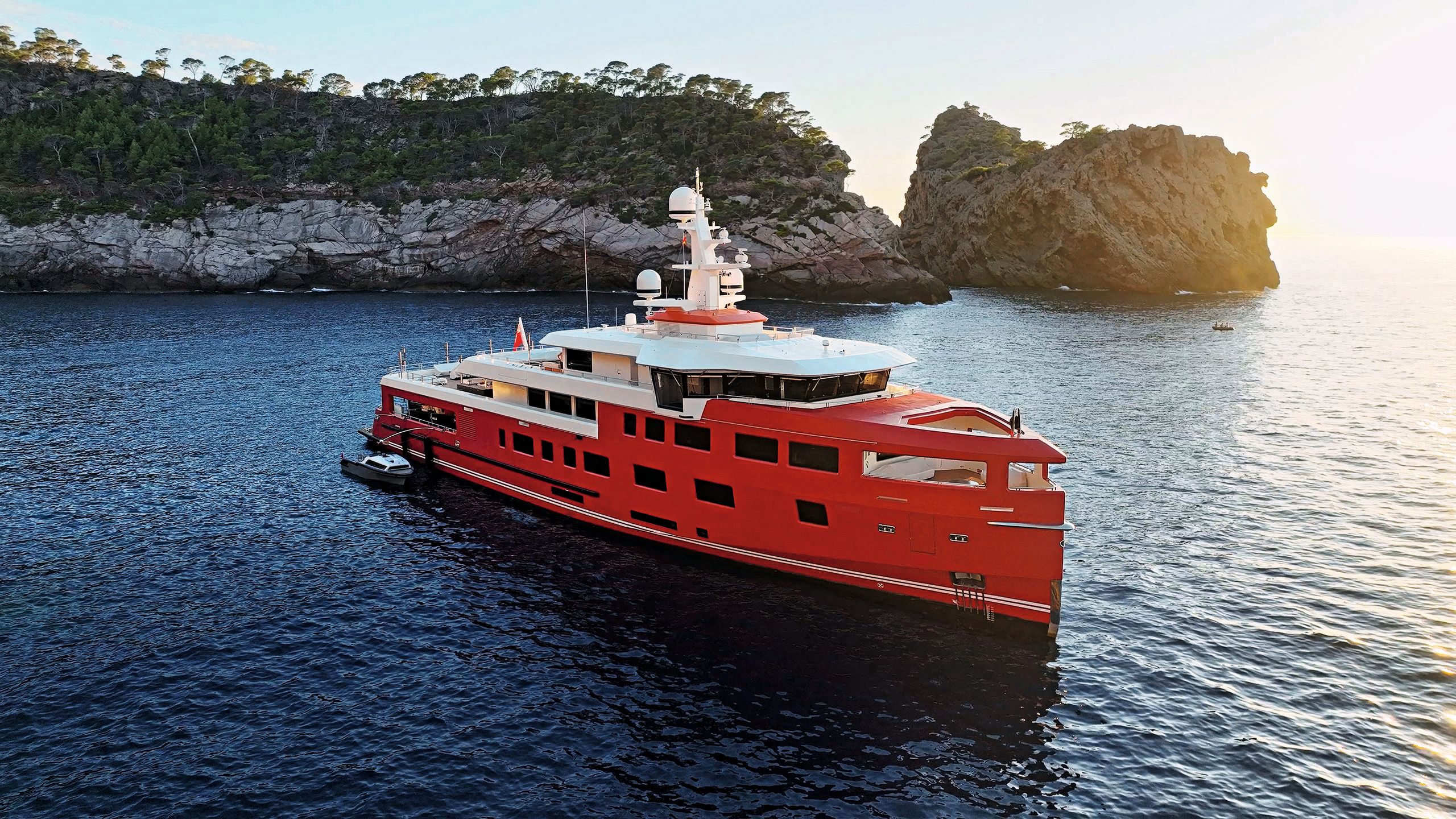 Wide shot of Akula on the water, with a rocky island behind with trees. She has bright red paintwork
