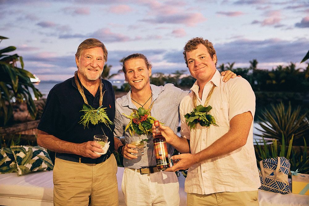 Stream has short grey hair and is wearing sunglasses and a navy blue Polo shirt. He has a necklace made of foliage around his neck and is standing next to a younger man in the middle and a middle-aged man to the right. Both are wearing short-sleeve shirts and trousers in light colours