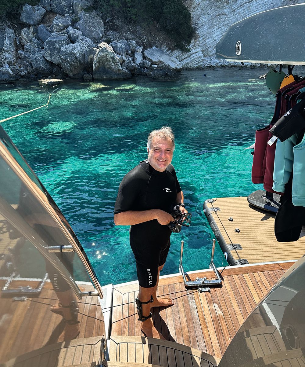 Apostolos is standing on the deck and the waters are turquoise behind him. He's wearing a black wet suit.