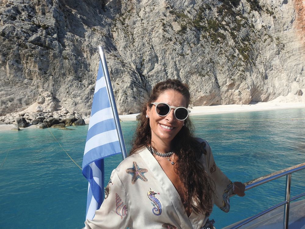 Ioanna Elena Markou has long brown hair and is wearing round sunglasses and a white top with a starfish and seahorse on. She is on the deck of the boat, smiling at the camera, and there is a Greek flag behind her.