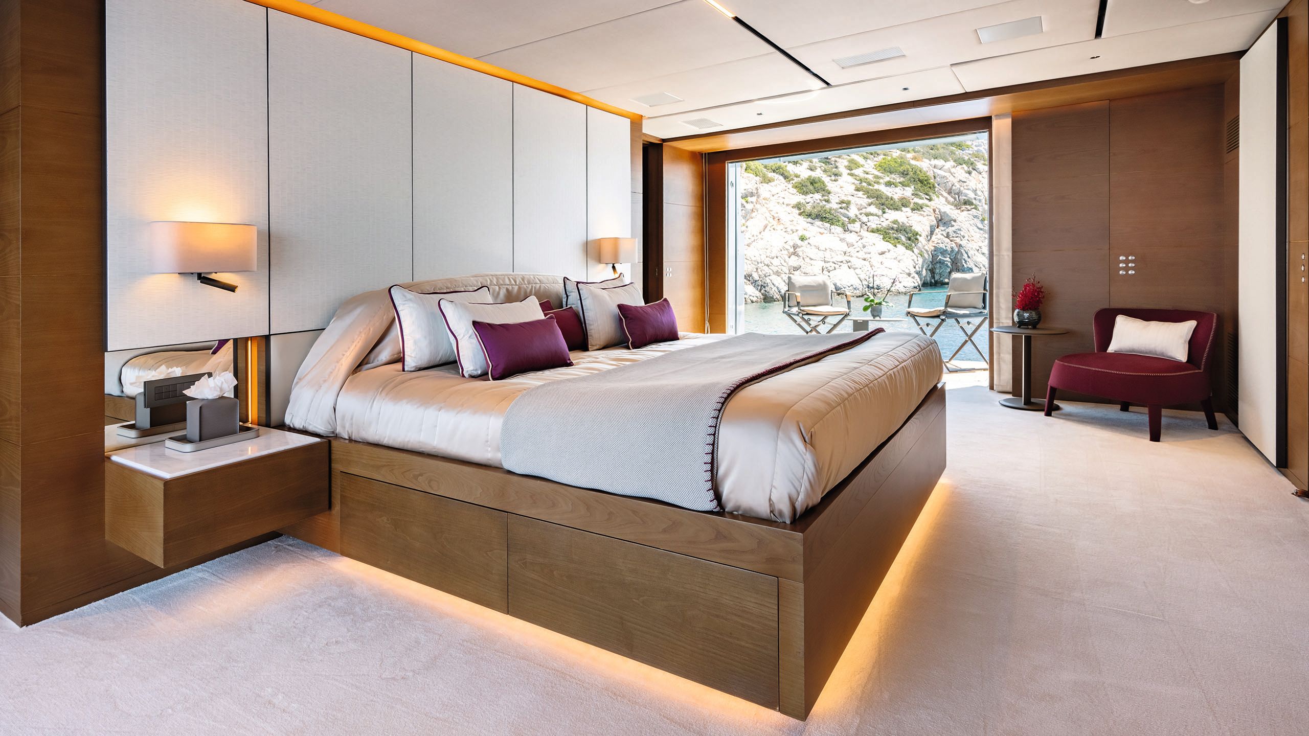 Master cabin with large bed in the centre. The floor is a neutral colour and the bed is under-lit. The furnishings are burgandy and neutral with darker wood surrounds.