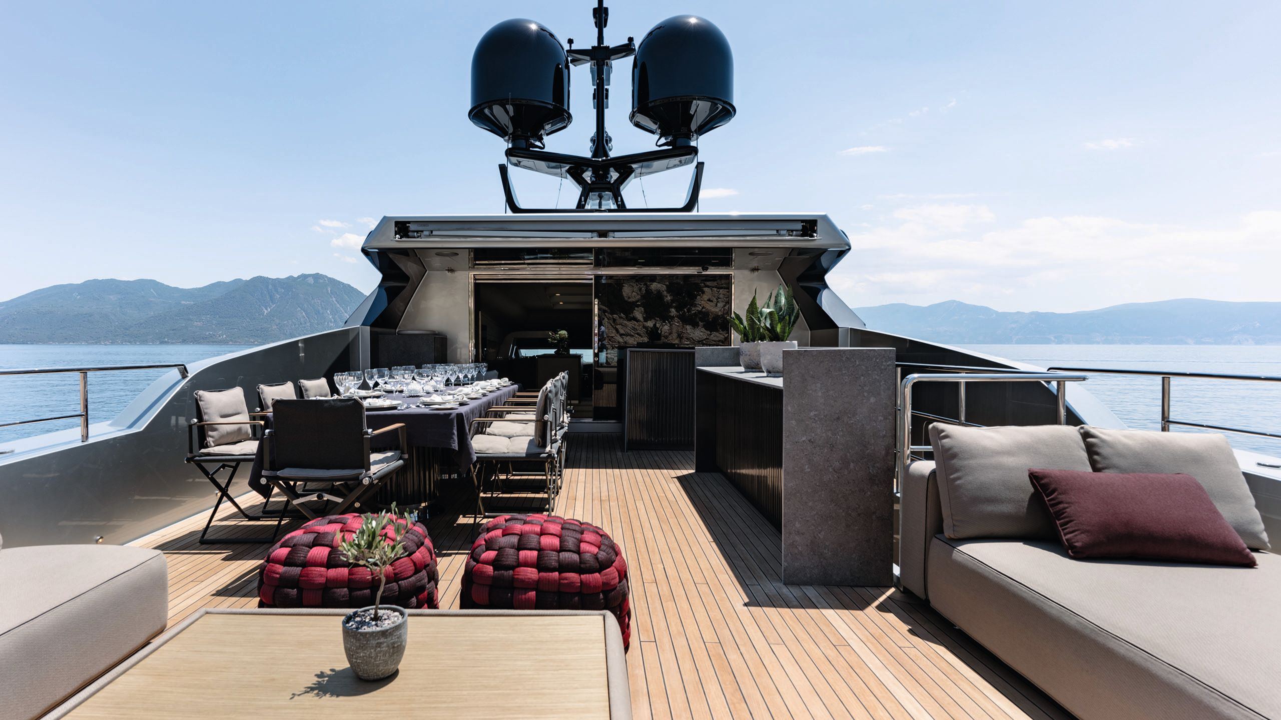 A wide exterior shot of a deck with a long dining table filled with wine glasses and place settings. In the forefront are two soft pouffs that are red and burgandy check, and a grey sofa to the right