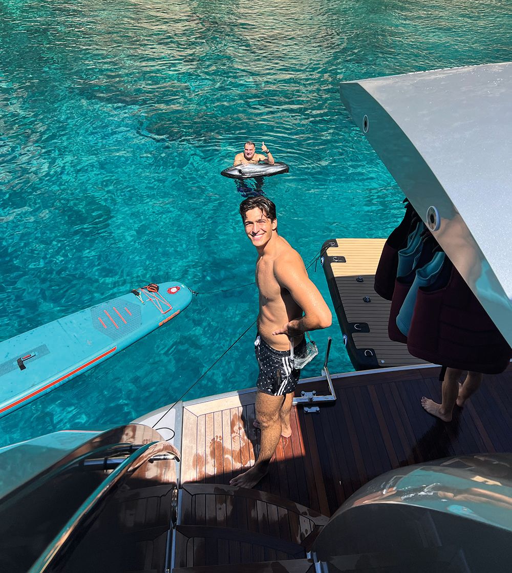 Tristan standing on the side of the boat with Apostolos in the turquoise water behind