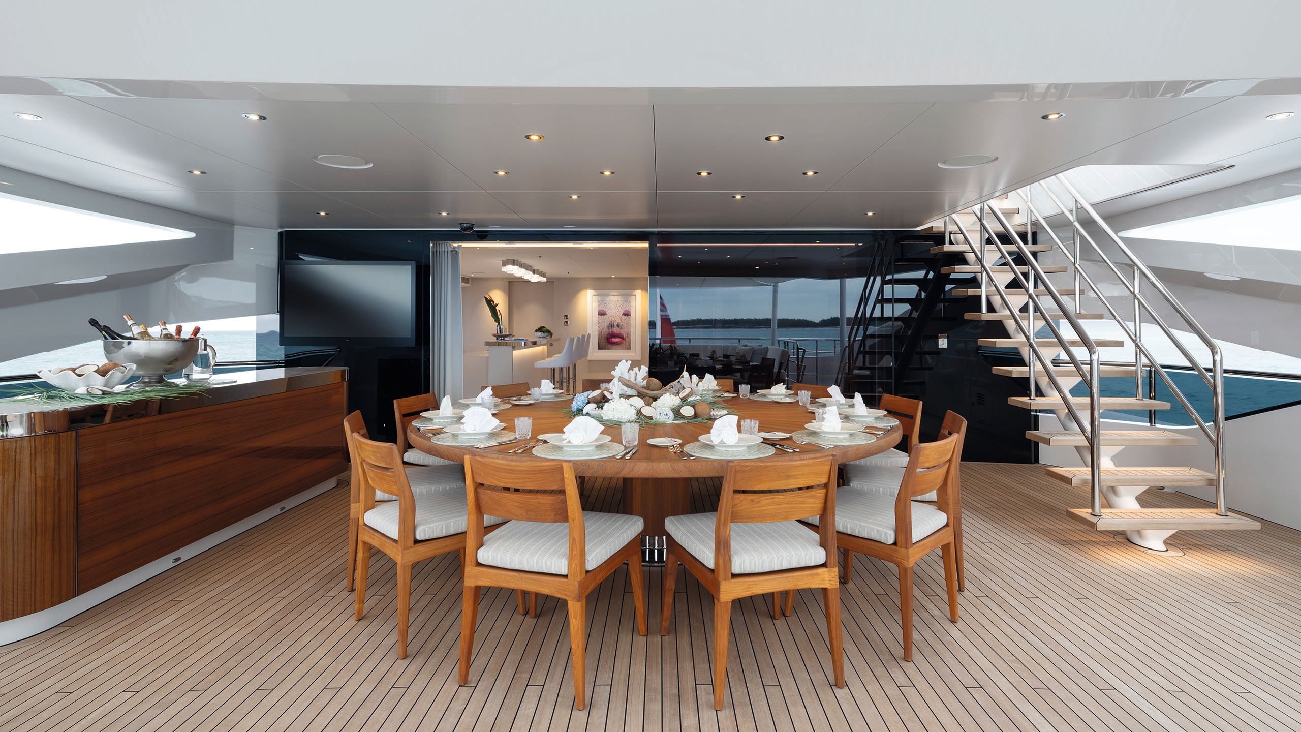 A circula dining table on the deck in a teak colour with matching chairs and grey and white striped cushion pads