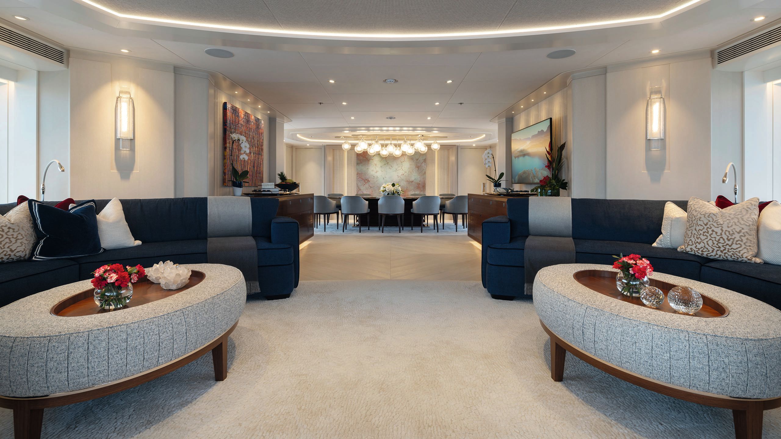 Wide shot of the main saloon which has sofas in a navy blue colour and oval coffee tables with grey upholstering. In the background is the dining table and chairs with a chandelier above it