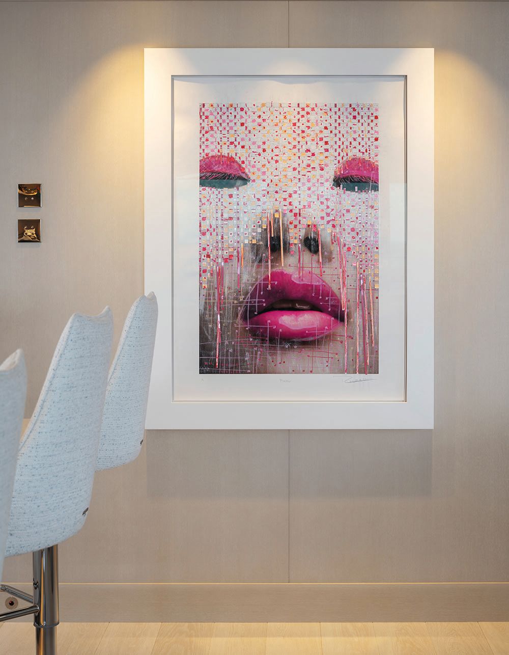 Artwork on the wall in a white frame and mount. It shows the close-up eyes, nose and lips of a face; the eyes and lips are hot pink