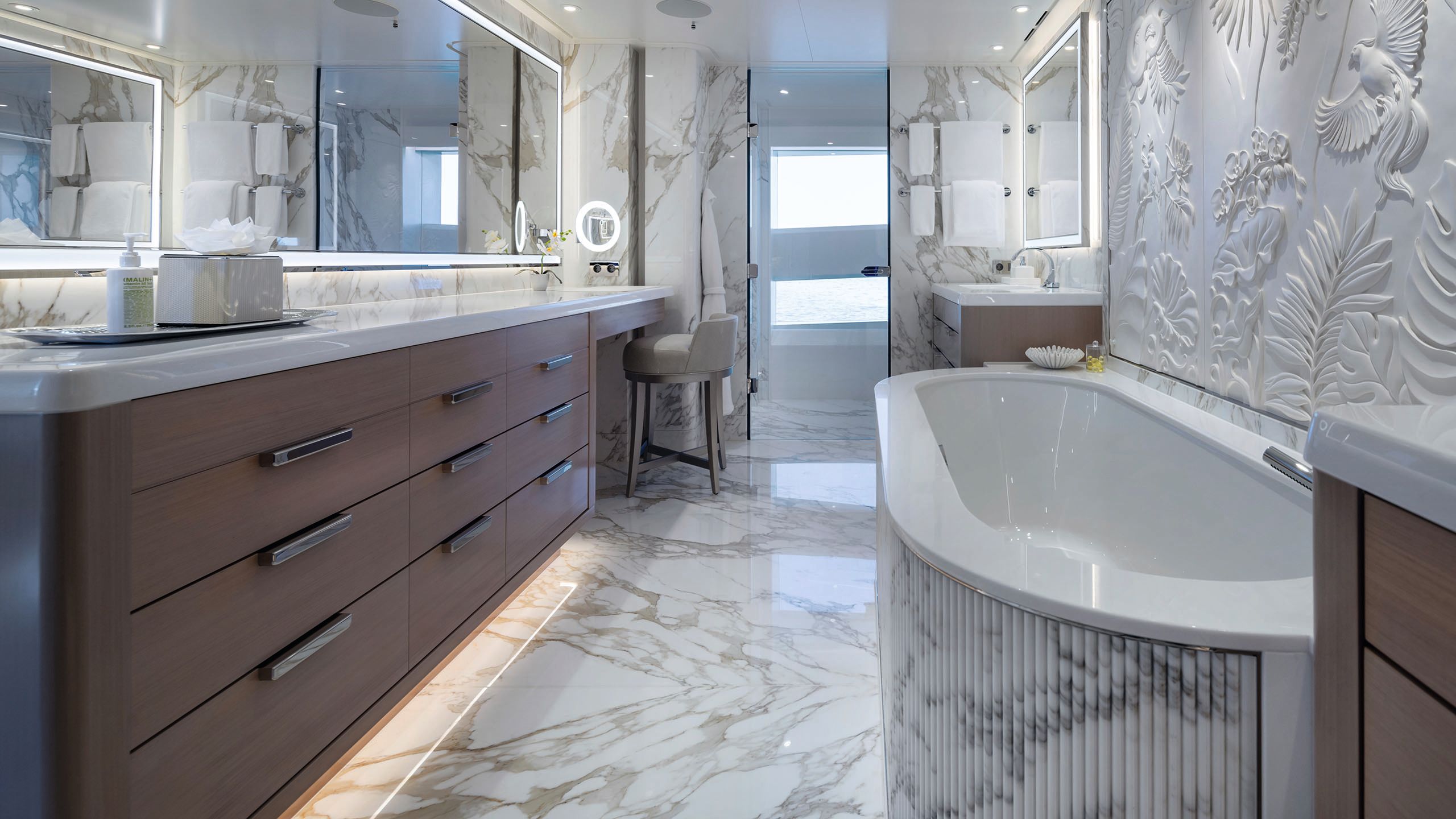 Wide shot of the bathroom - to the left is a bank of dark wood drawers and a marple top and mirror above. Opposite is a white bath with the plaster sculpture on the wall next to it