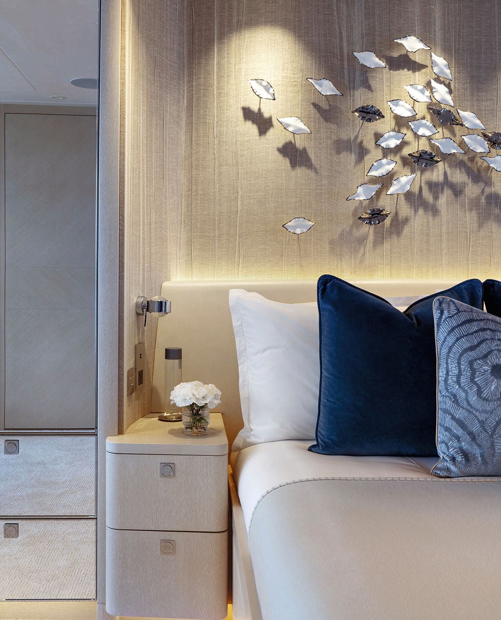 Partial view of the side of a bed in neutral colours with blue pillows. Above it is a leaf sculpture