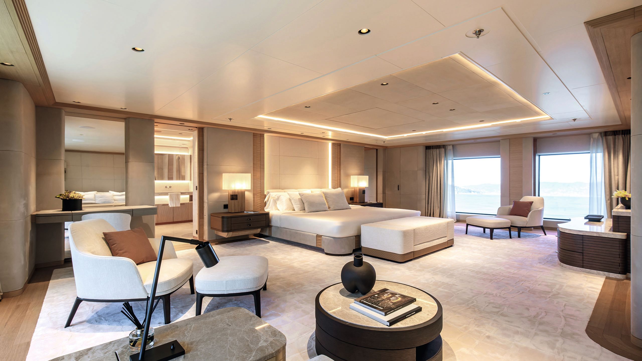 Large bedroom suite with bed at the centre. It is decorated in neutral colours with brown accents. In addition the bed, there are two armchairs and footstools and a bench at the foot of the bed