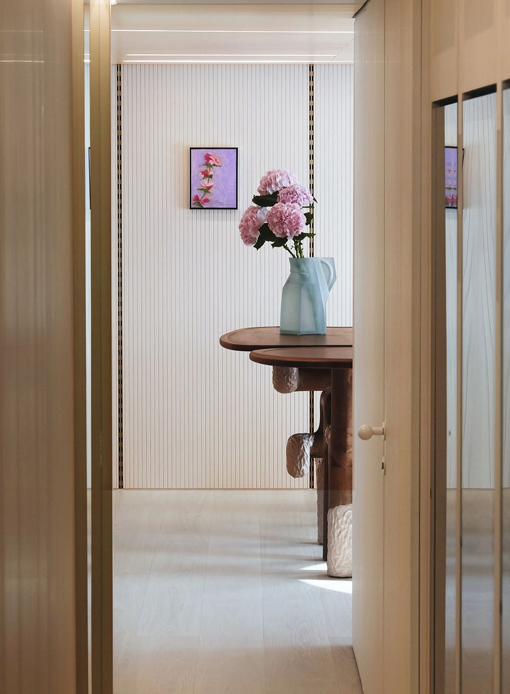 View looking down a hall into a room which has ribbed walls and a purple and pink picture of a flower. There is a dark-wood table and an abstract blue jug on it, holding pink flowers with large blooms