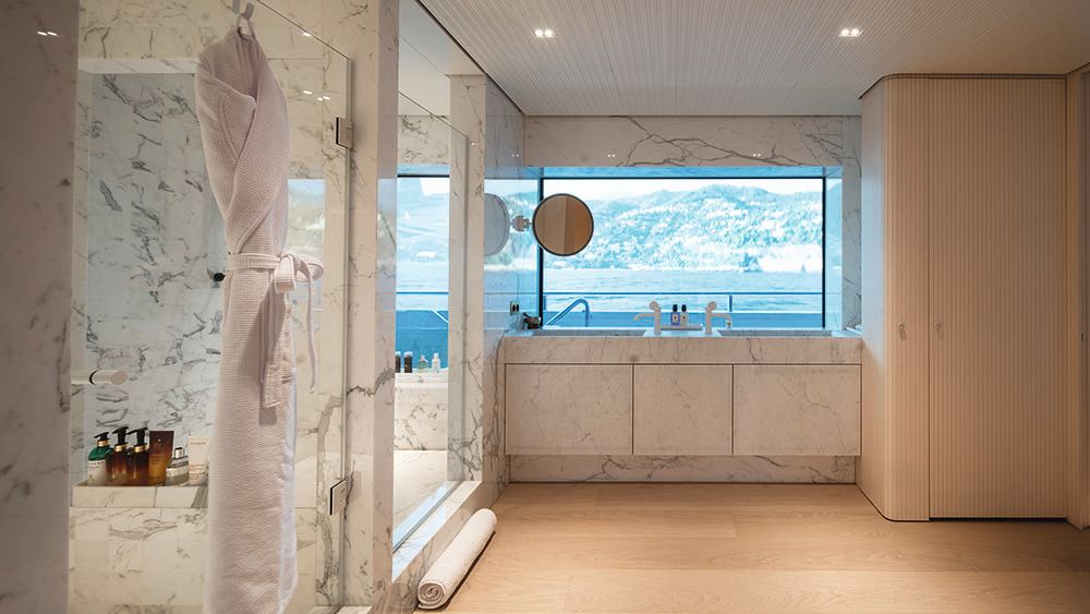 Wide shot of the en suite, which is made from white marble. A large vanity the width of the window looks out on the sea. A dressing gown is hanging on the shower door in the foreground