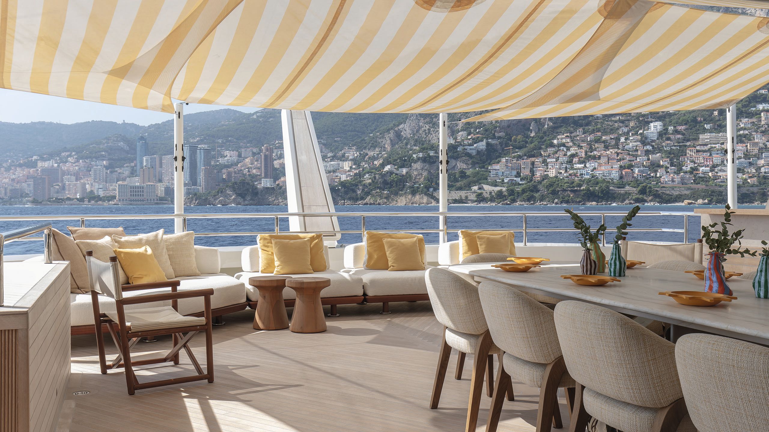 Deck area covered with the same white and yellow striped awning. There is a large long dining table in the centre and large sunpads in cream behind with yellow cushions. There is a folding chair in teak with cream material.