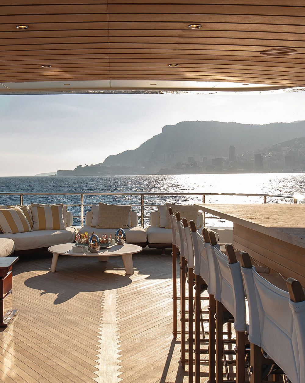 View of the deck which has a silver railing around it. There is an outdoor bar area with high chairs in teak and white. In the background are sunpad-style soft seats with yellow and white scatter cushions