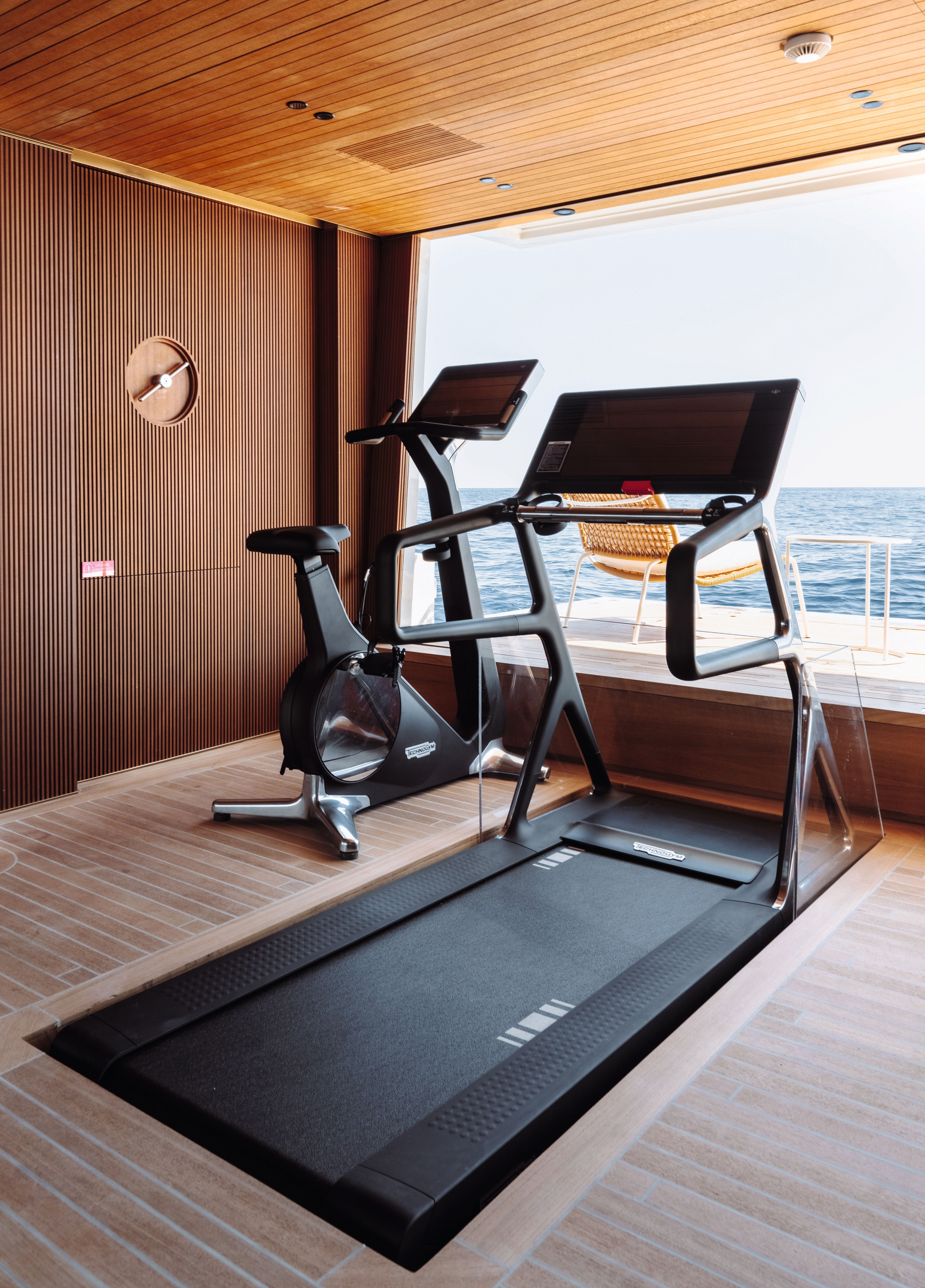 Side view of a treadmill facing a large window looking out on to the sea. There is ribbed panelling on the walls and a minimalist clock on the wall