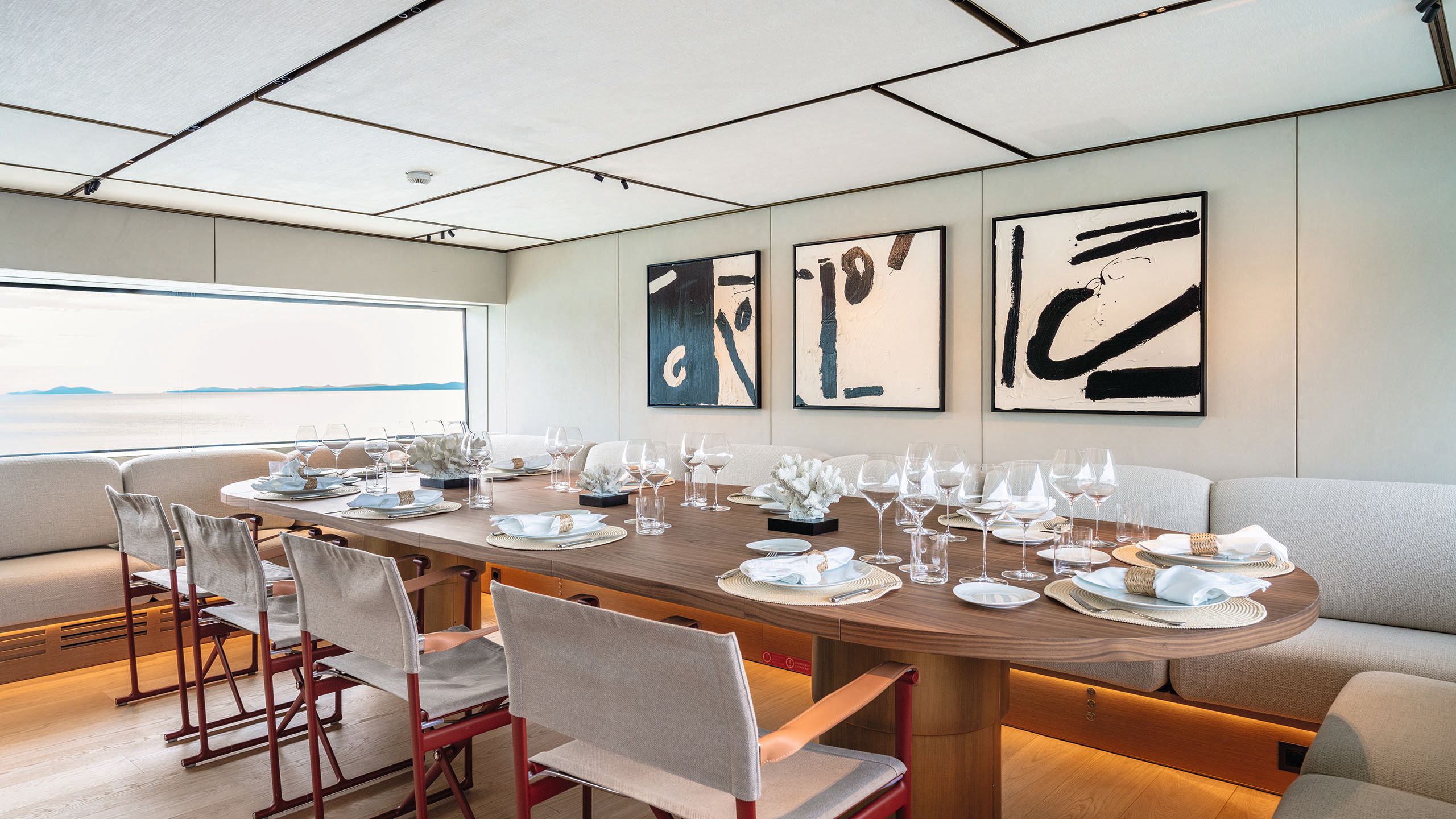 Looking into the media lounge from a different angle. There is abstract white and black art on the walls, a table with geometric white bases and white L-shaped sofas. A large table with extra chairs is set in front of the sofa