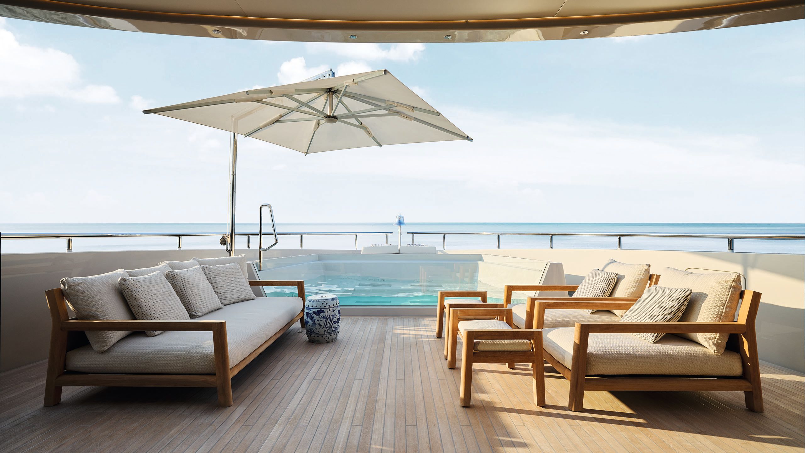 Outdoor deck with sofa and arm chairs with foot rests. A parasol is providing shade and at the back is a small pool