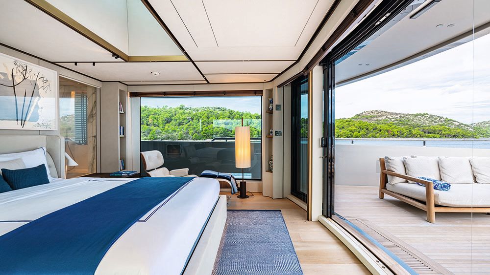 Side view of a bad made up with white linens and blue throw and pillows. It faces a large window which opens on to the deck