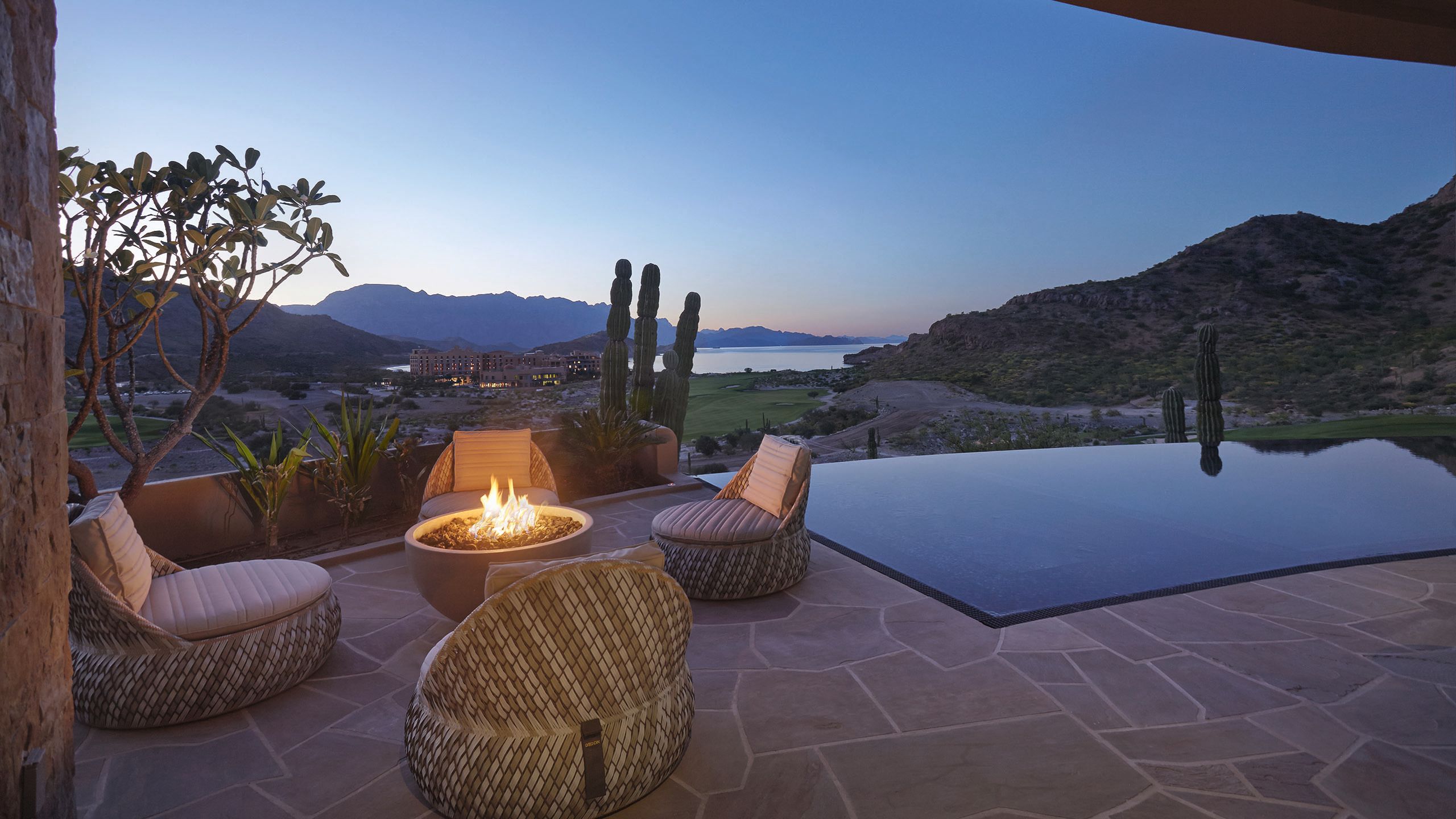 A patio area at dusk with rattan-style comfy chairs in front of a circular firepit. There is cacti surrounding the edges and mountains in the distance