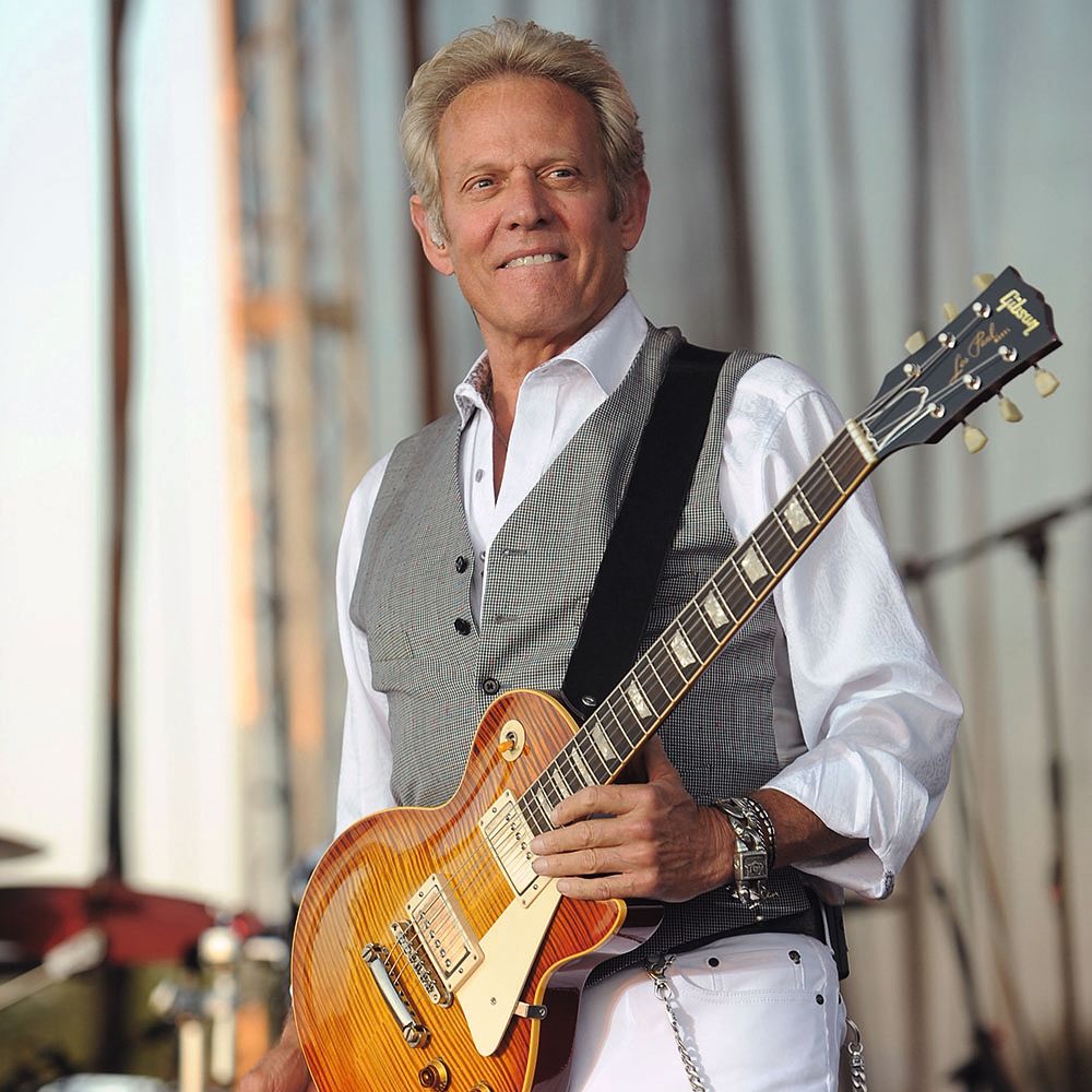 A man with grey hair wearing an electric guitar, white shirt, grey waistcoat and white trousers