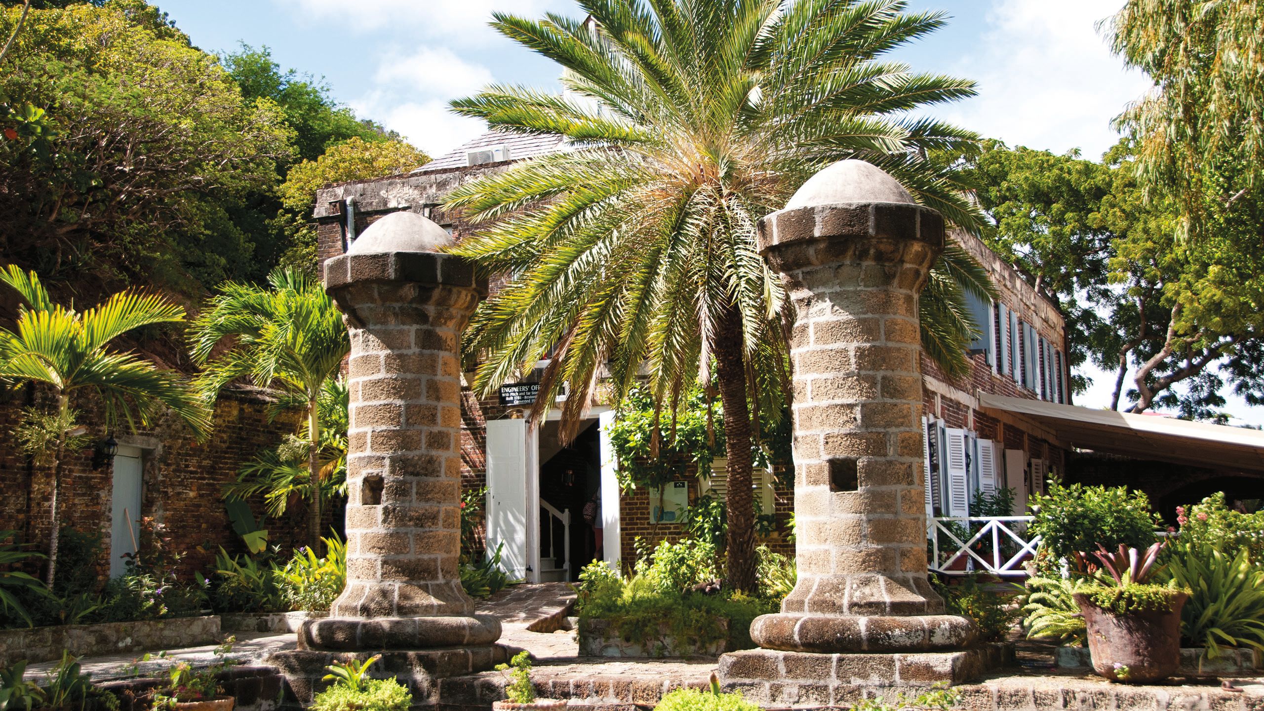 Two large brick pillars at the front of a colonial-style house, surrounded by palm trees