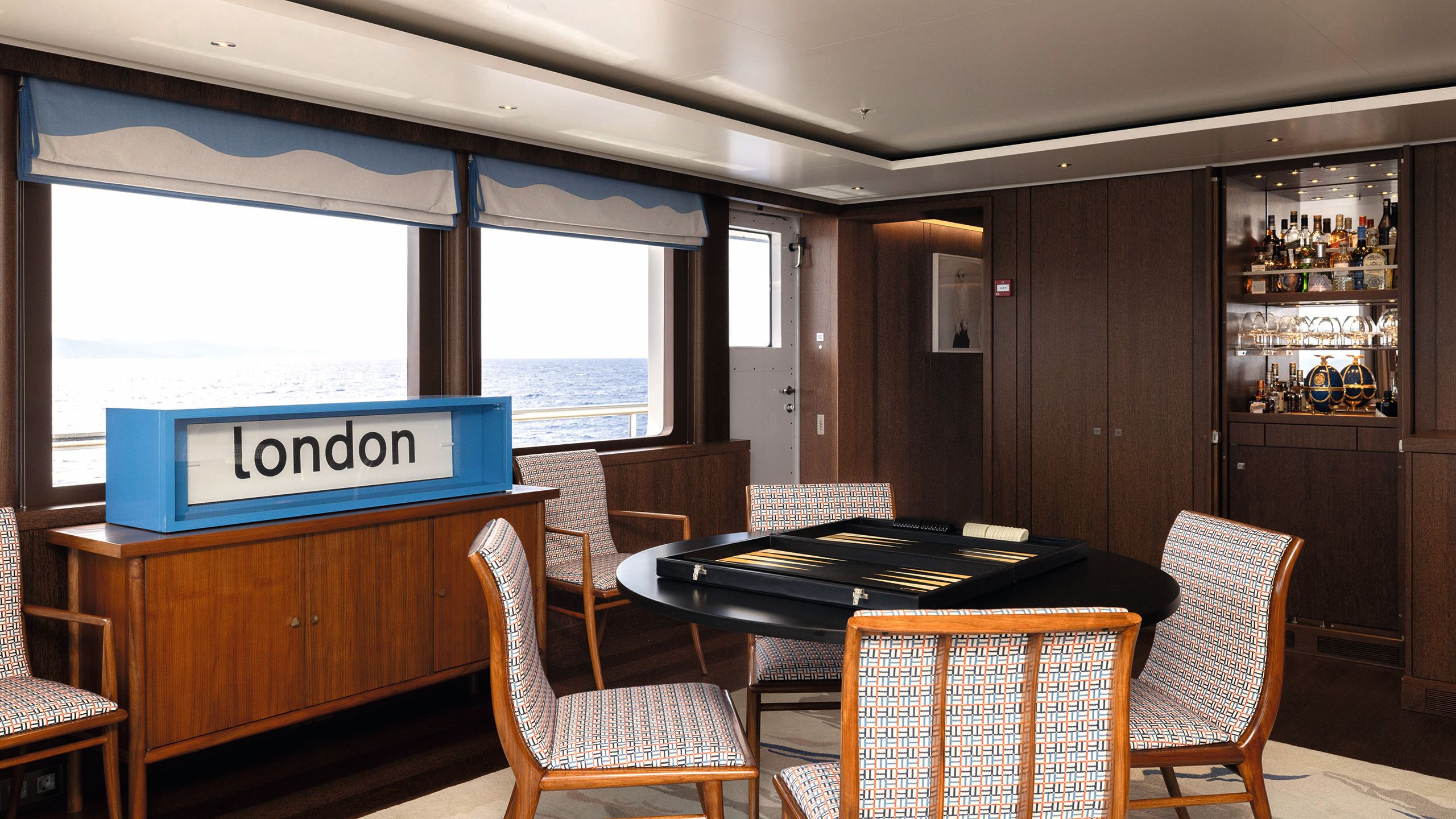 Round black table with four chairs and a backgammon set on top. There is a large blue box with a white sign that says London in black letters