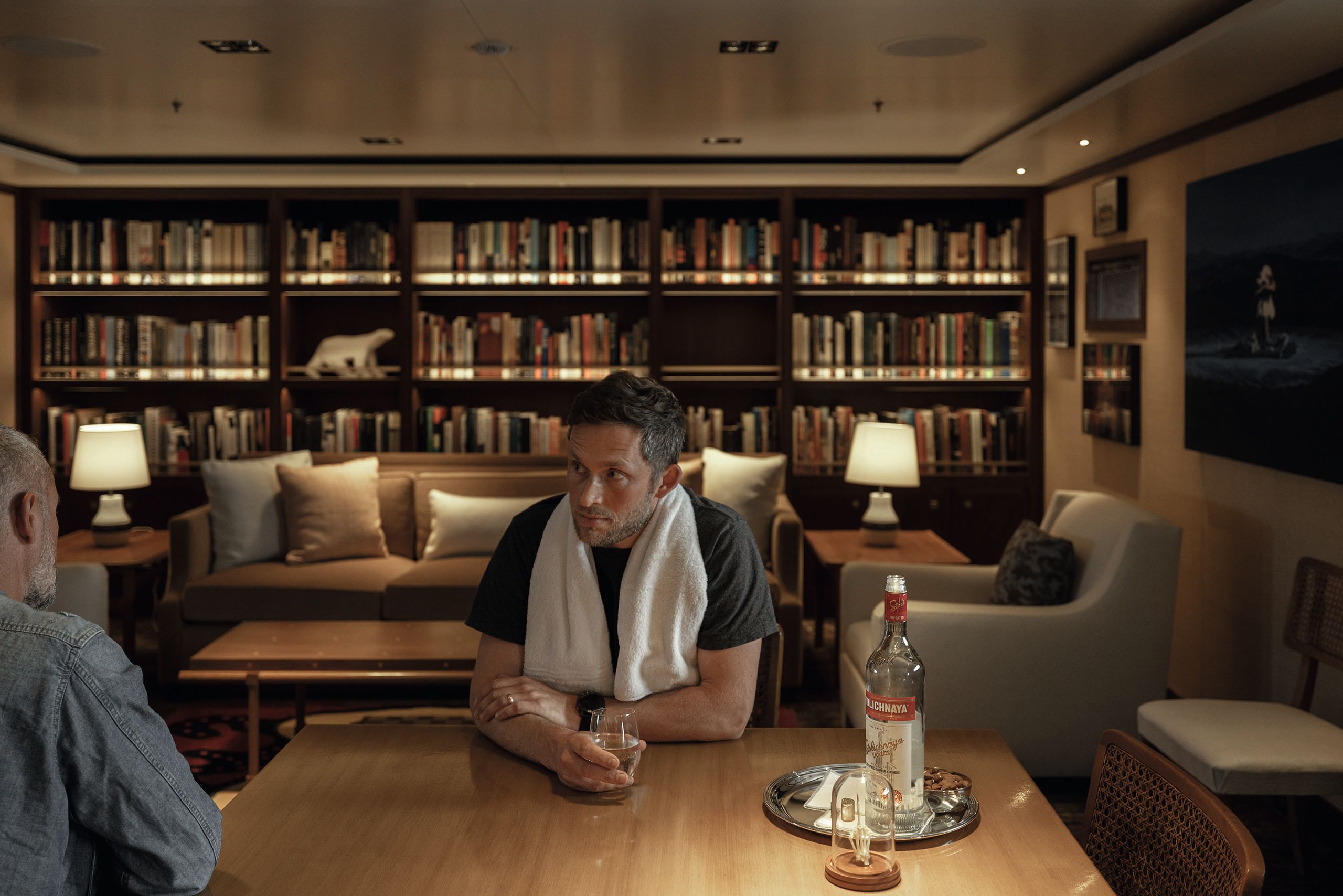 A man sitting at a table in the library holding a drink. He has a towel around his neck. There are shelves filled with books behind him