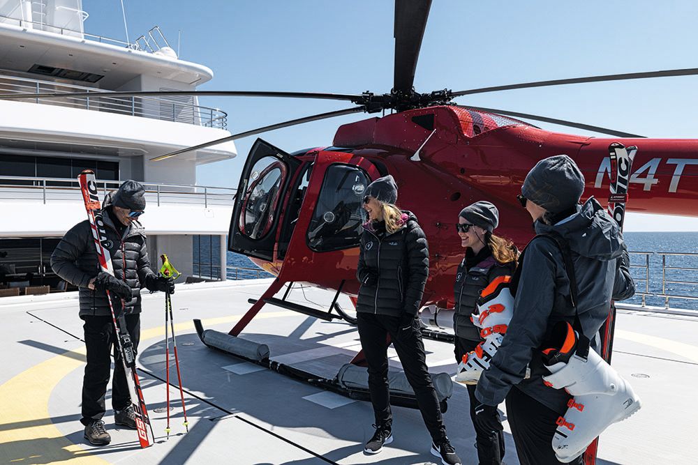 Group of people standing in front of the red helicopter. One man is holding a set of skis